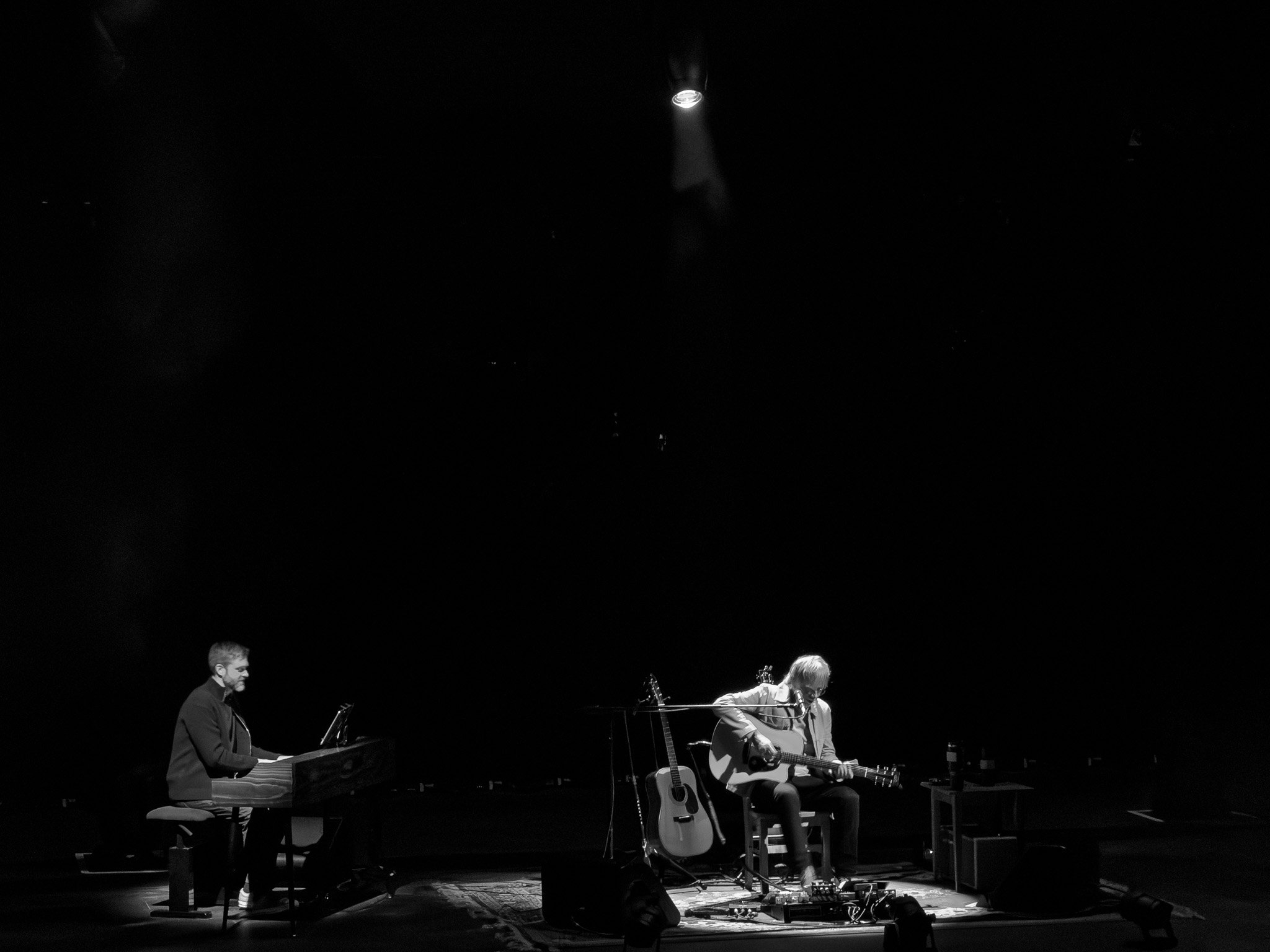 Two musicians perform on stage: one playing piano and the other playing guitar and singing into a microphone, under a spotlight.
