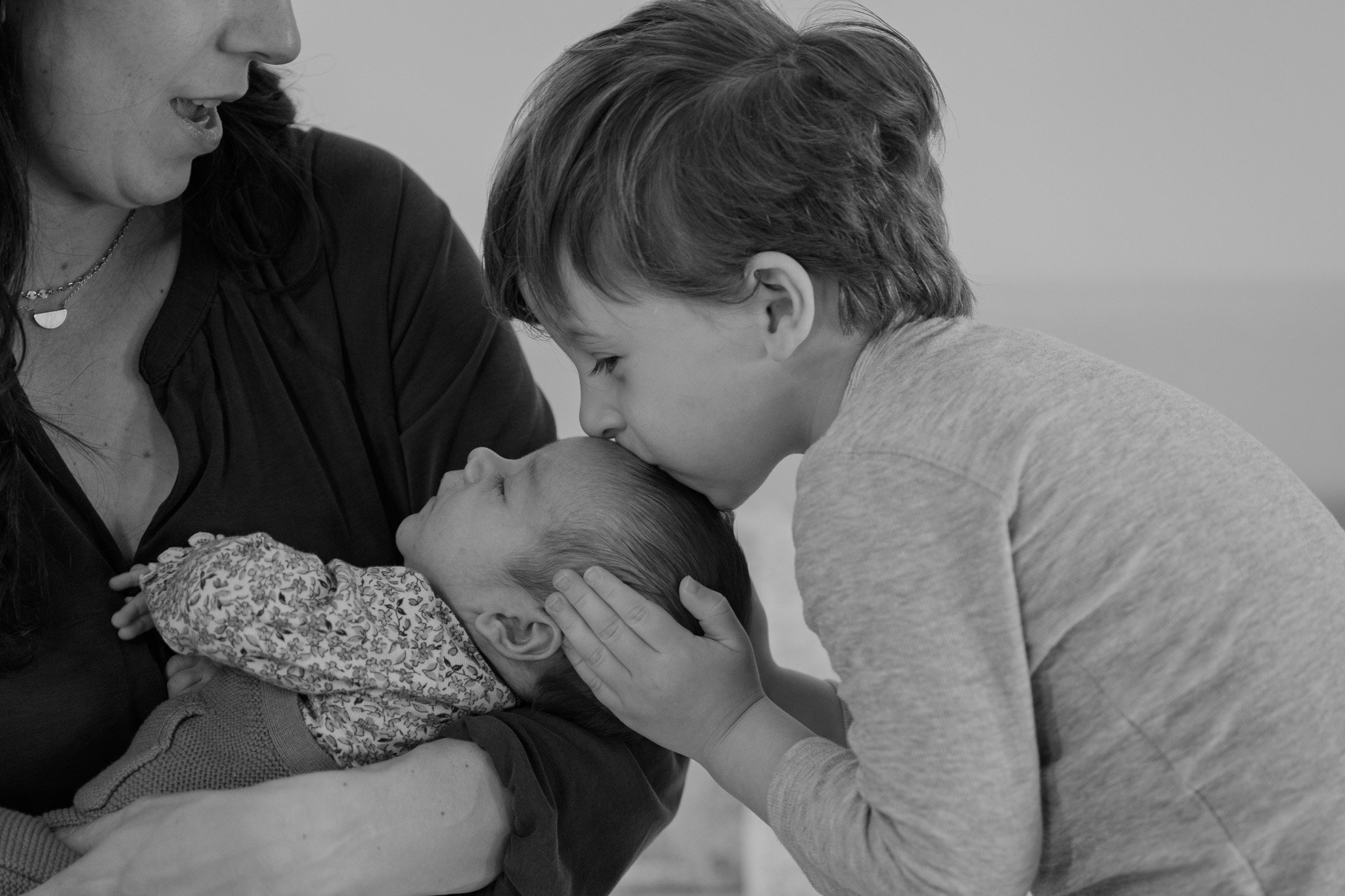 A woman and a young boy gently kissing their baby sibling on the forehead in black and white.