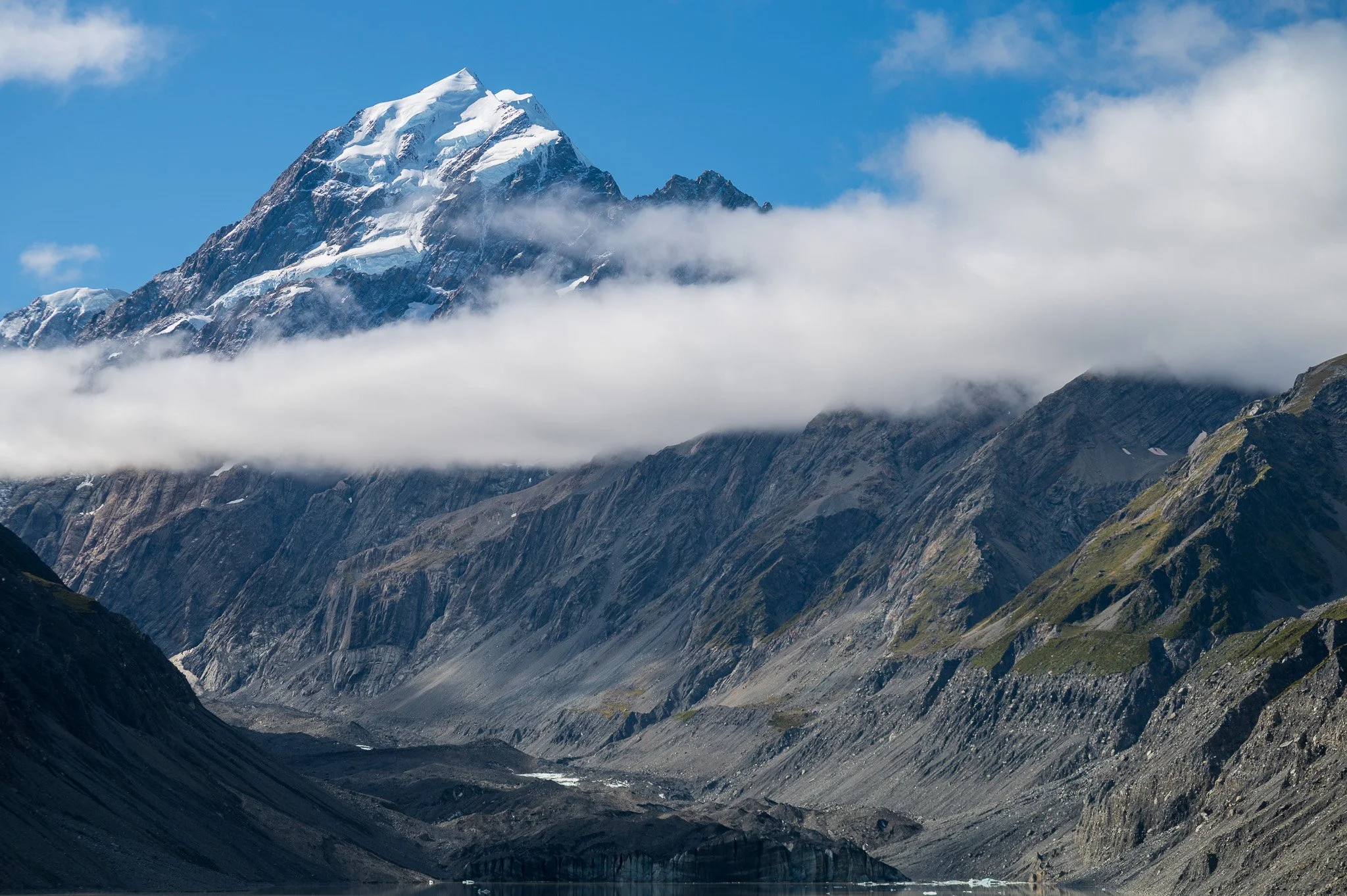 Snow-capped mountain peak rising above clouds with rugged rocky slopes in the foreground.