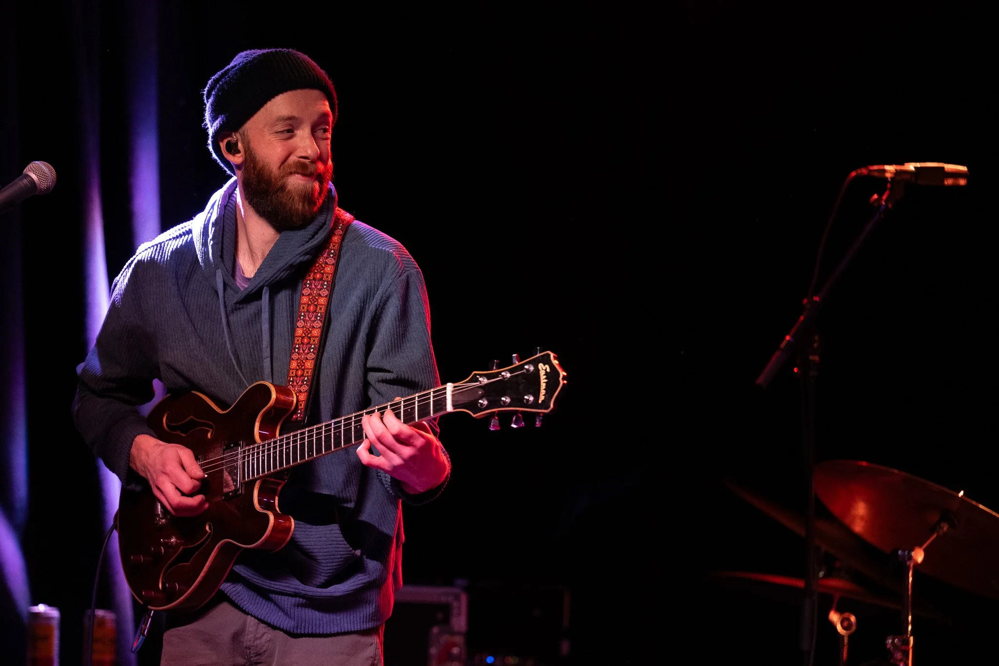 A man with a beard wearing a black beanie and a blue jacket playing an electric guitar on stage, with a microphone and music stand nearby, against a dark background.