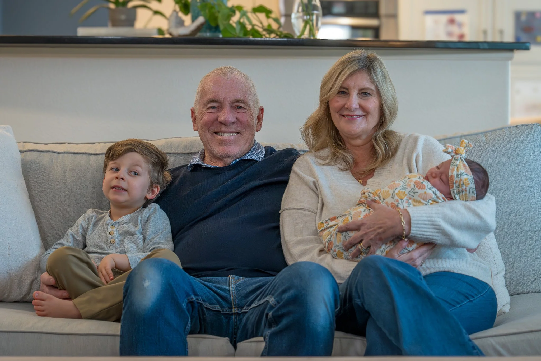 A family of four sitting on a beige couch in a living room. The grandfather is smiling, the grandmother is holding a sleeping baby girl wrapped in a floral blanket, and a young boy is sitting beside them, looking slightly to the side.