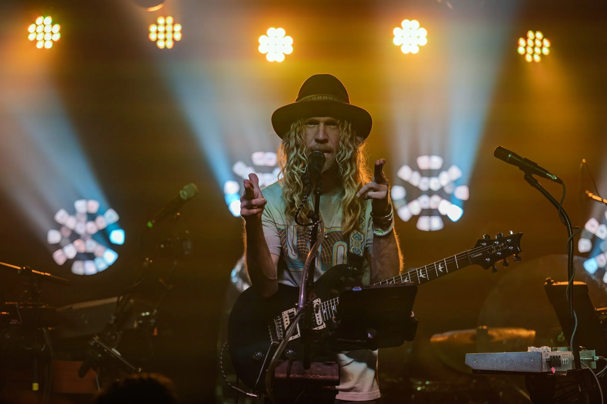A musician with long curly hair, wearing a hat, performing on stage with a guitar. The stage is lit with bright, colorful lights in the background.