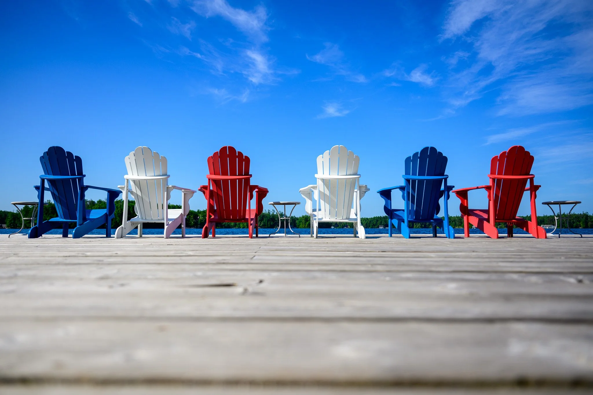 Colorful Adirondack chairs on a wooden dock by a lake under a clear blue sky. Muskoka, Canada.