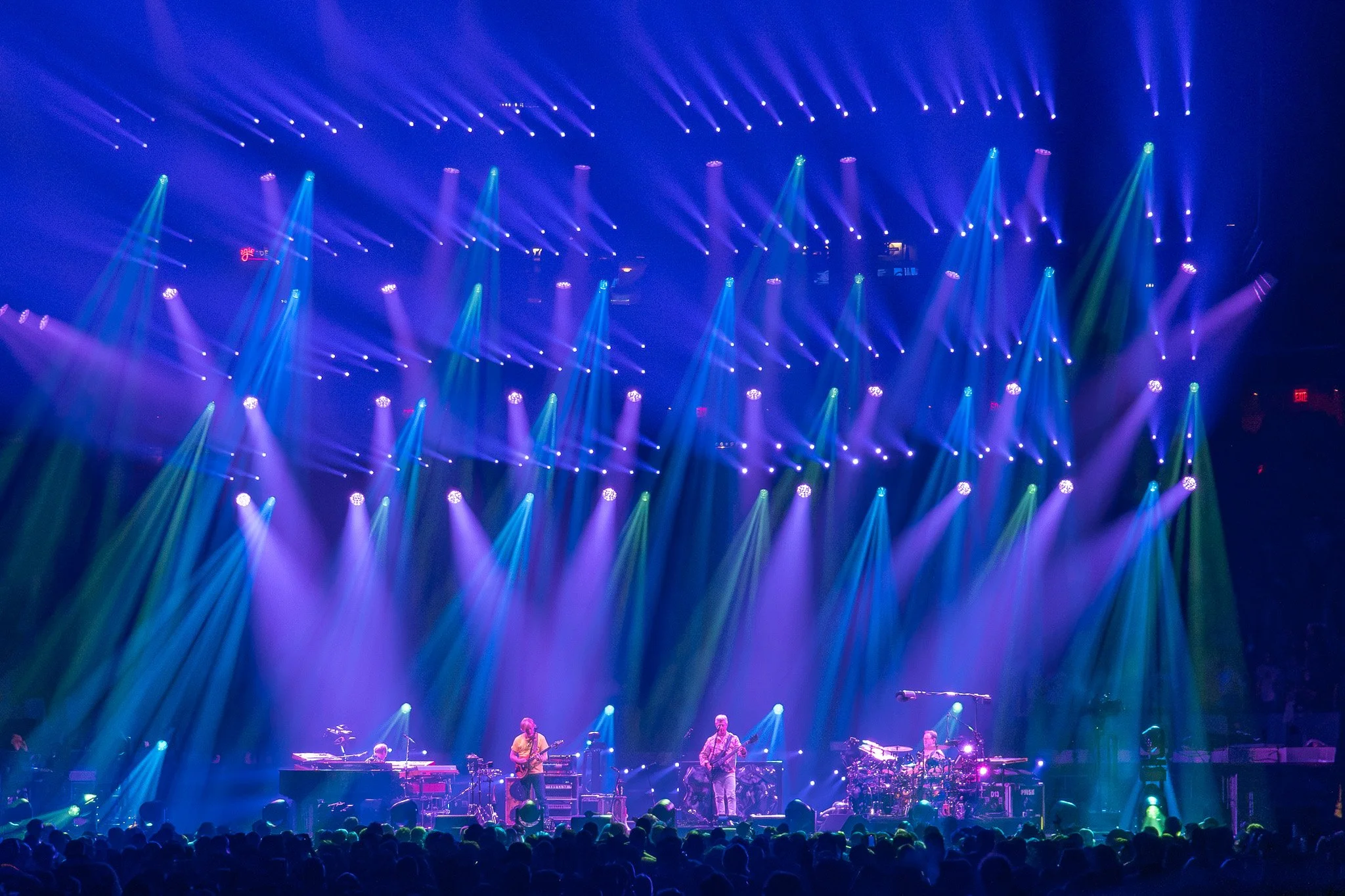A concert stage with a band playing under colorful stage lights in a dark venue filled with an audience.