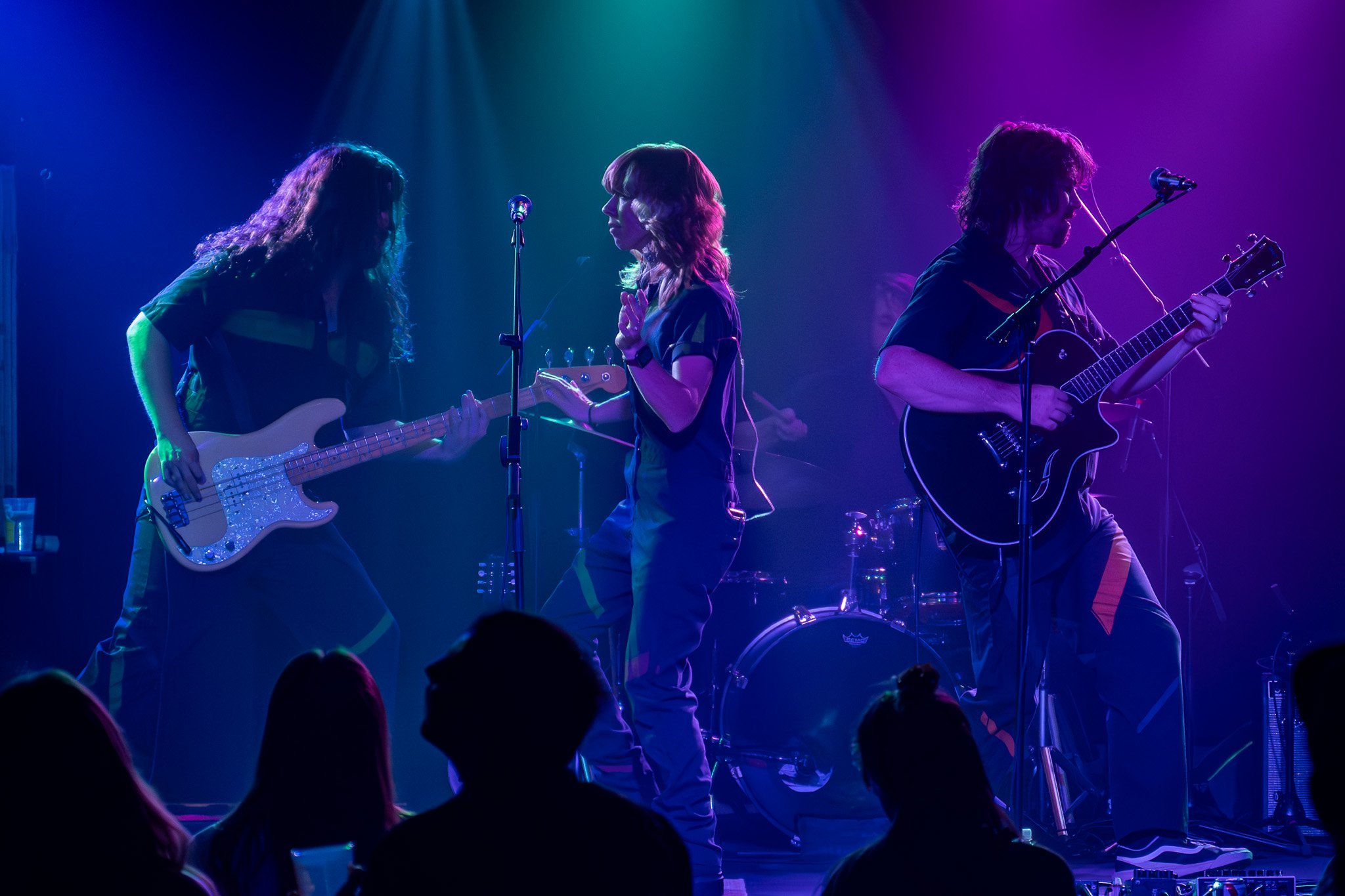 Three musicians on stage performing under colorful lights, one playing a bass guitar, another a keyboard, and the third singing into a microphone.
