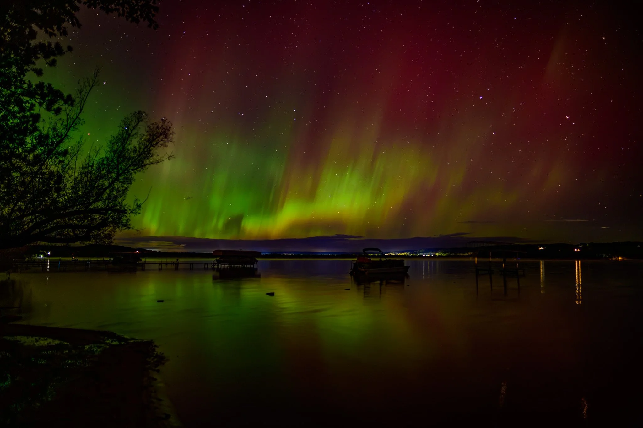 Northern lights illuminating the night sky over a lake with trees and boats in the foreground.