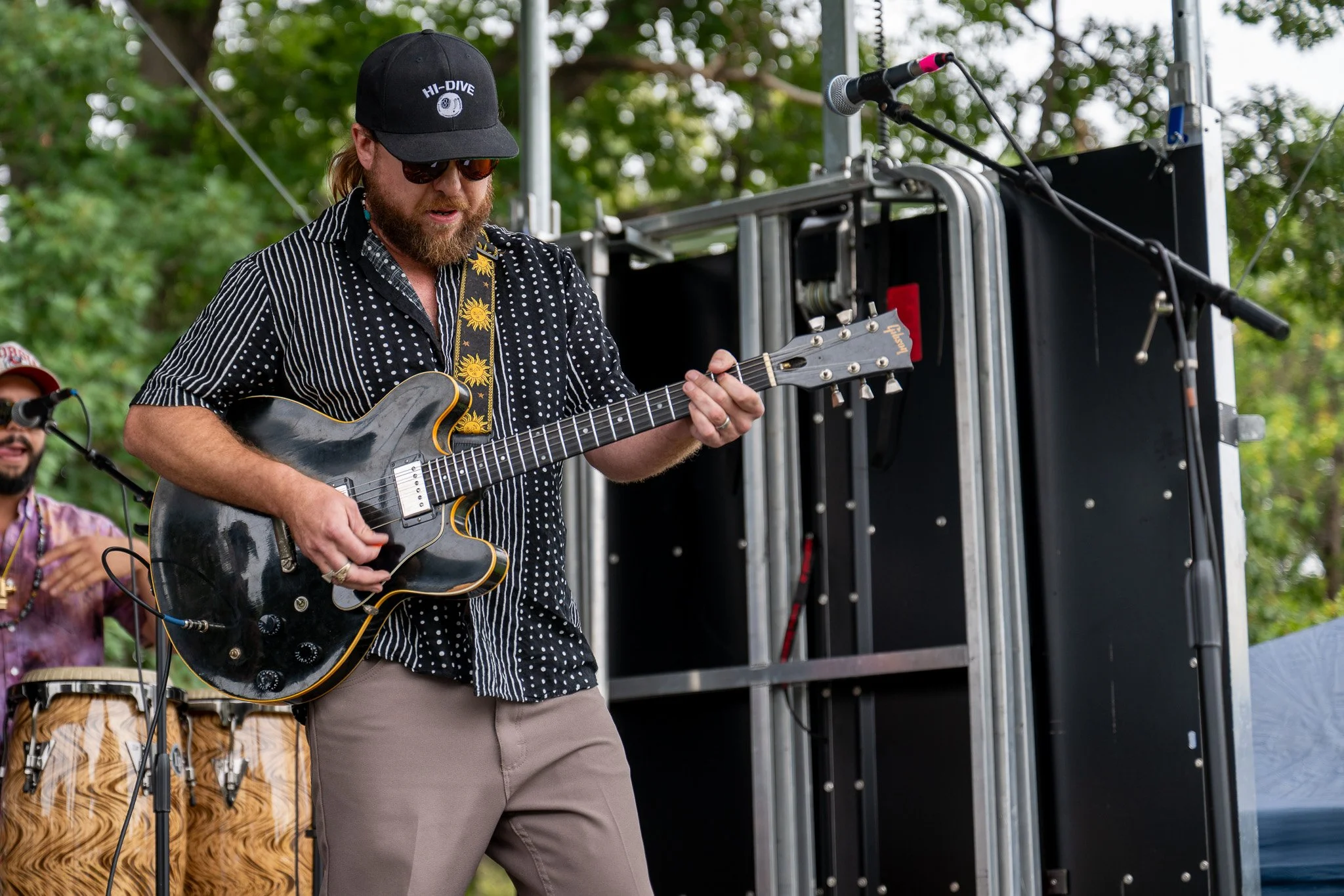A man with a beard, wearing sunglasses, a black cap with 'HI-DIVE' embroidered on it, and a black and white striped shirt, playing an electric guitar on a stage outdoors during daytime. There are trees in the background.