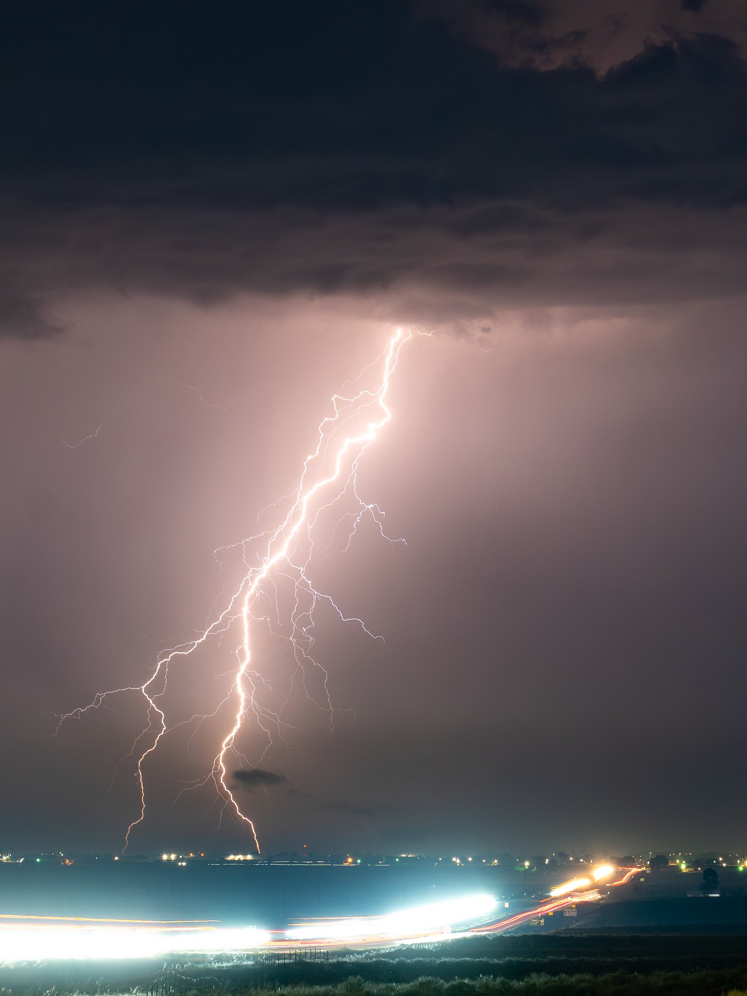 Lightning storm over a body of water at night, with dark clouds and bright streaks of lightning in the sky, and blurred car lights on the road below.
