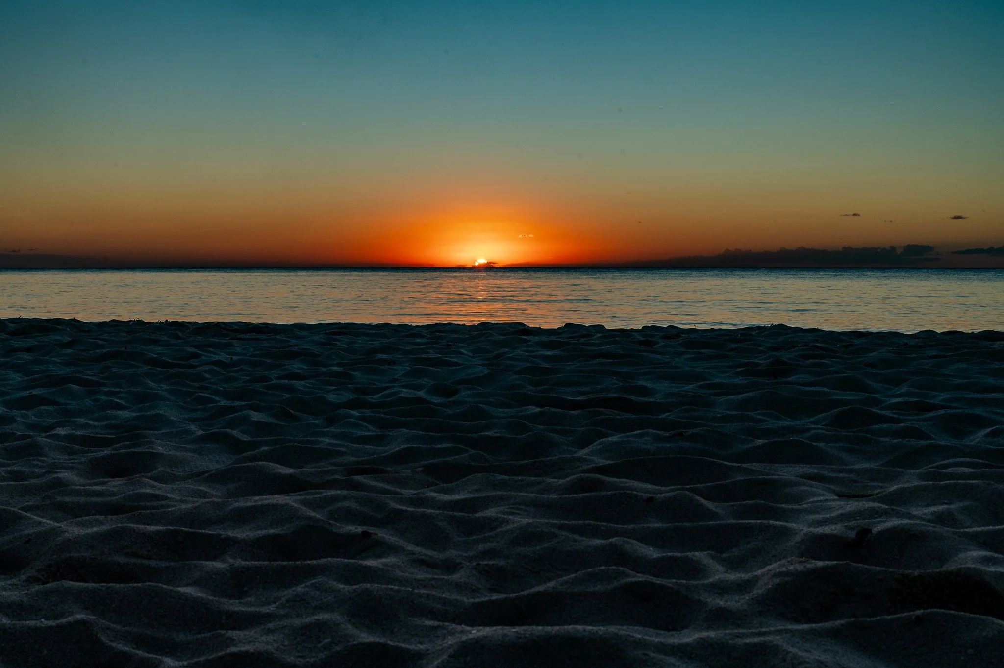 Sunset over calm ocean with sandy beach in the foreground.
