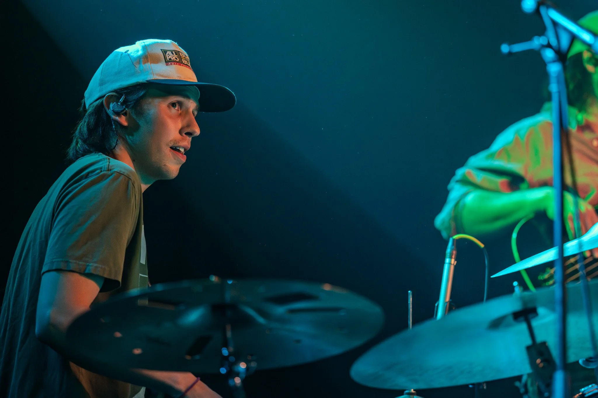 A musician with long hair wearing a beige shirt and a baseball cap, performing on stage with a drum kit, illuminated by colorful stage lights.