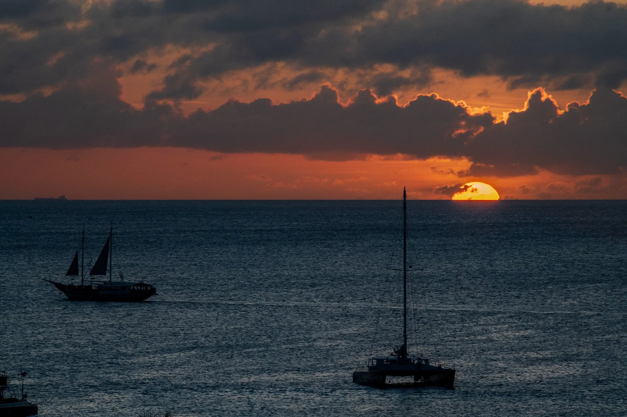 Sunset over the ocean with dark clouds, sailboats floating on the water, and the sun partially visible near the horizon.