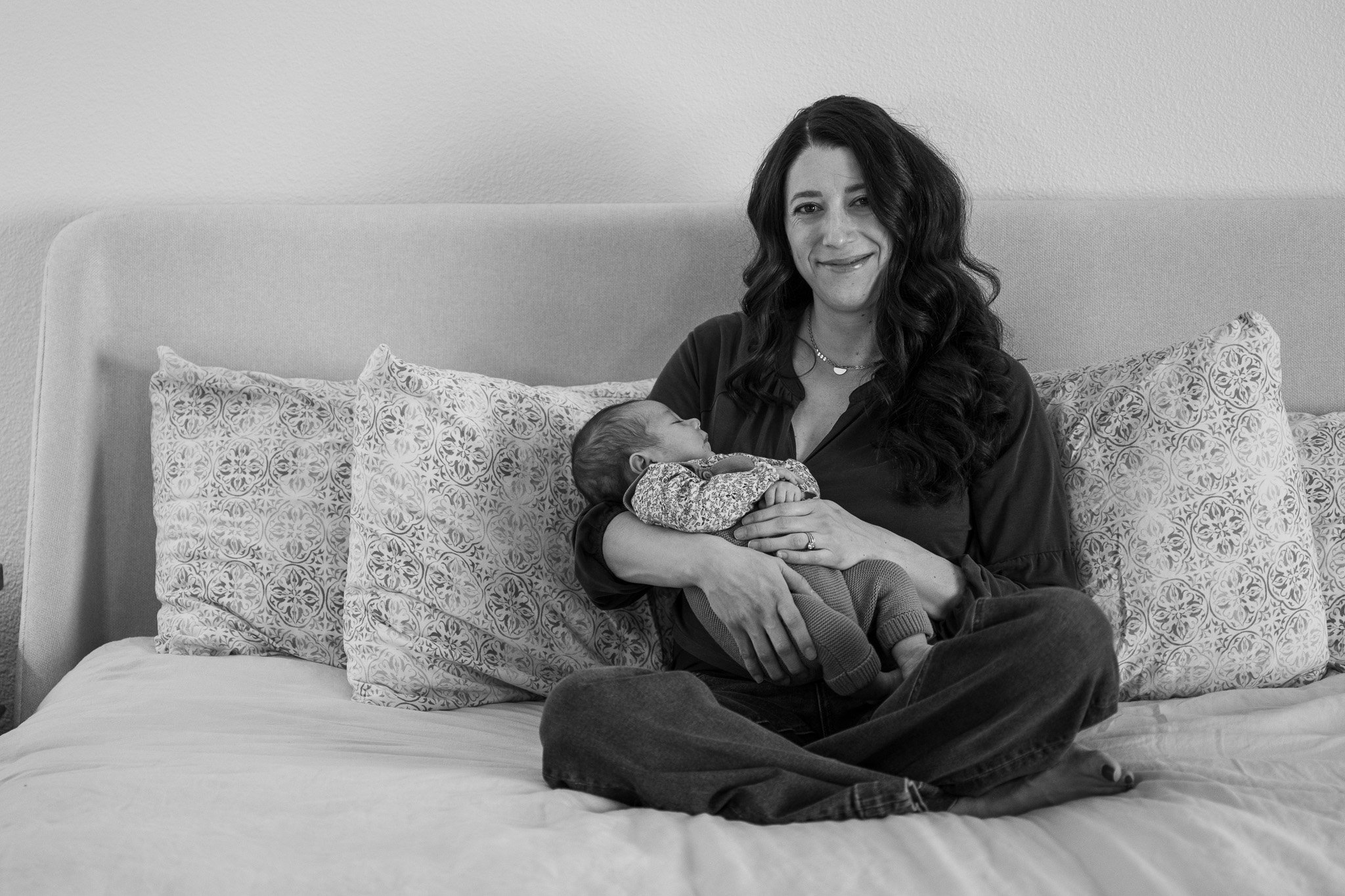 A woman with long, wavy hair sitting on a bed, holding a sleeping baby in her arms, smiling softly. The bed has patterned pillows and a plain headboard. The scene appears to be in a bedroom.
