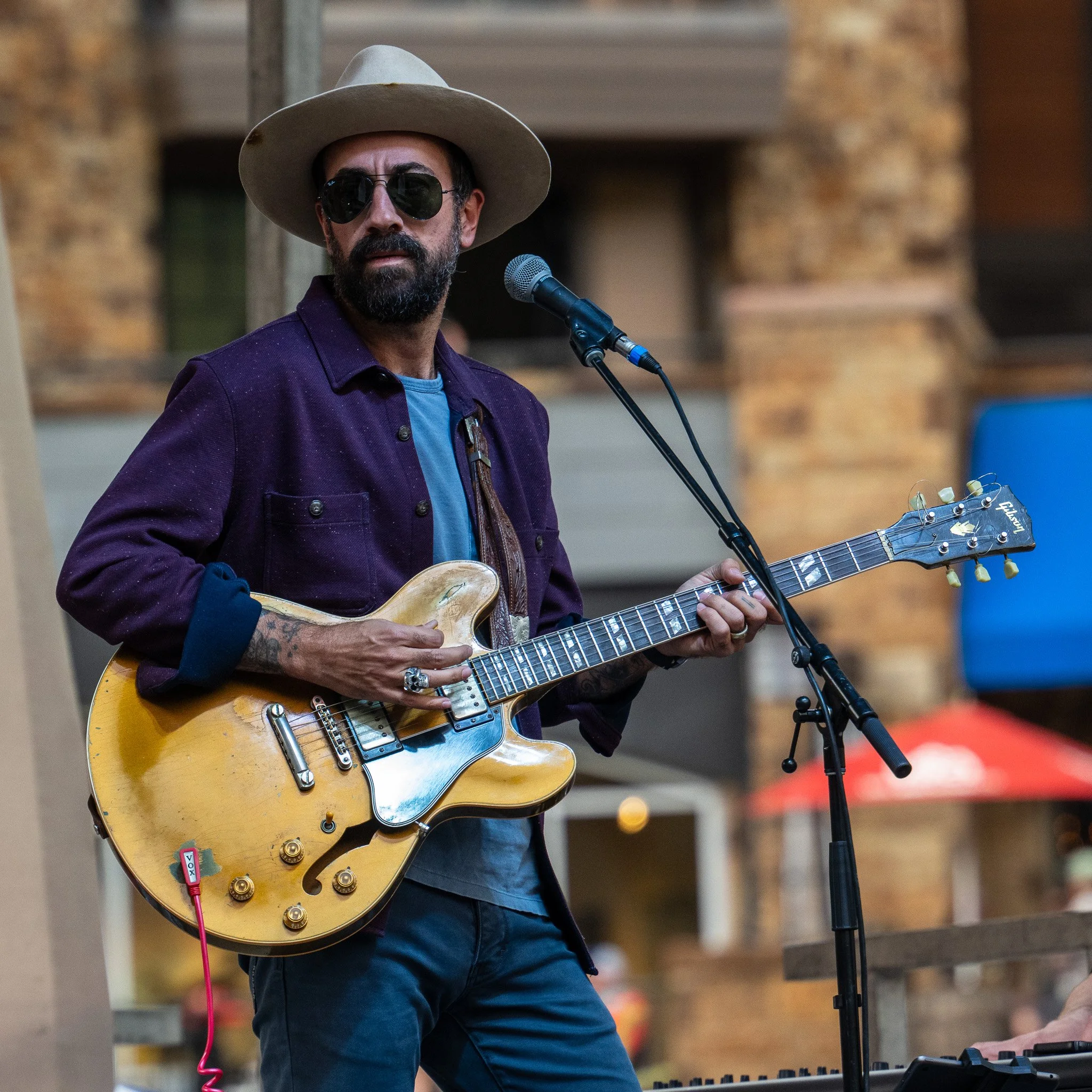 A man with a beard wearing sunglasses, a wide-brimmed hat, and a purple jacket playing an electric guitar and singing into a microphone.
