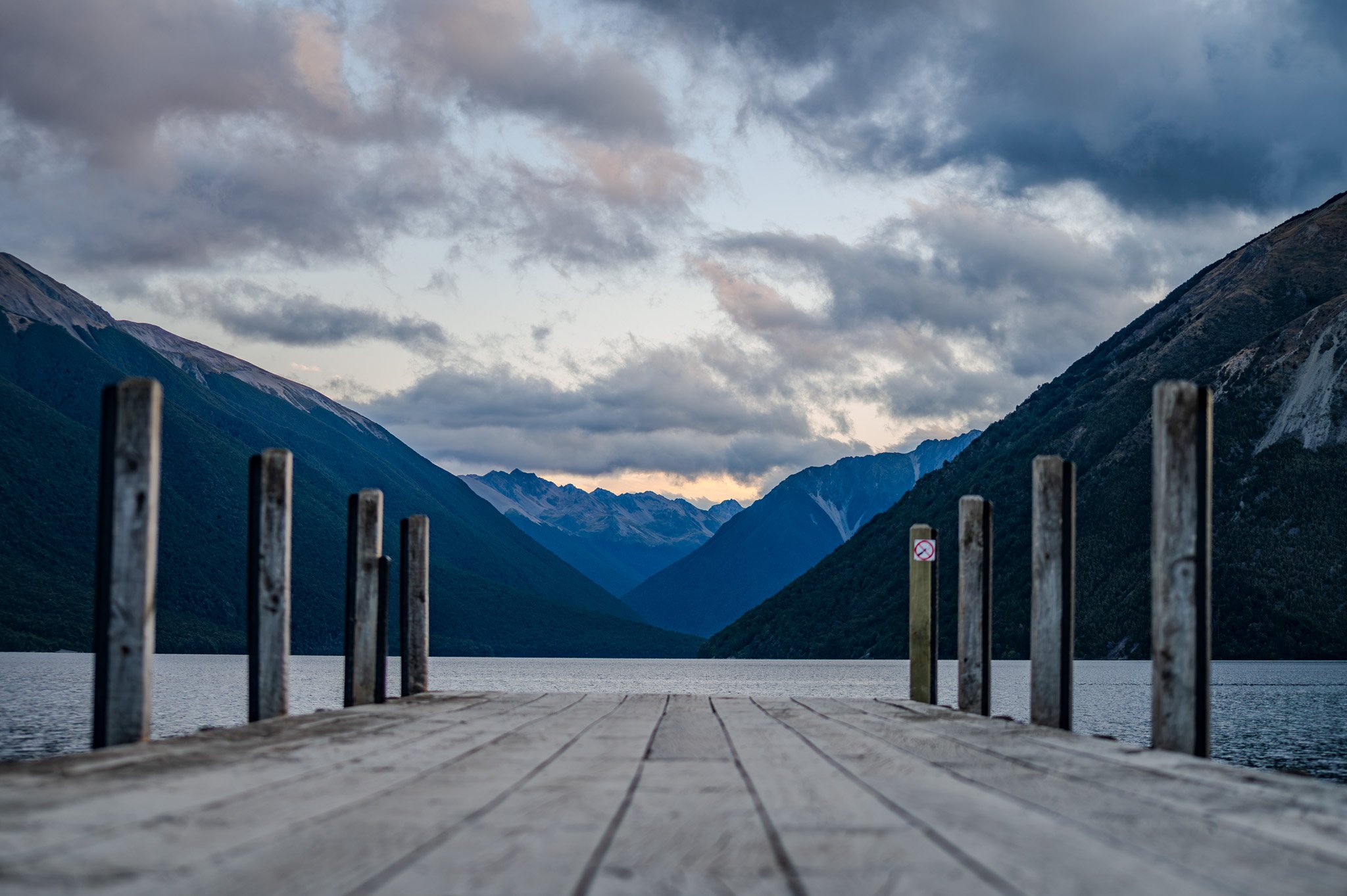 A wooden dock over a body of water with mountains in the background and cloudy sky above.