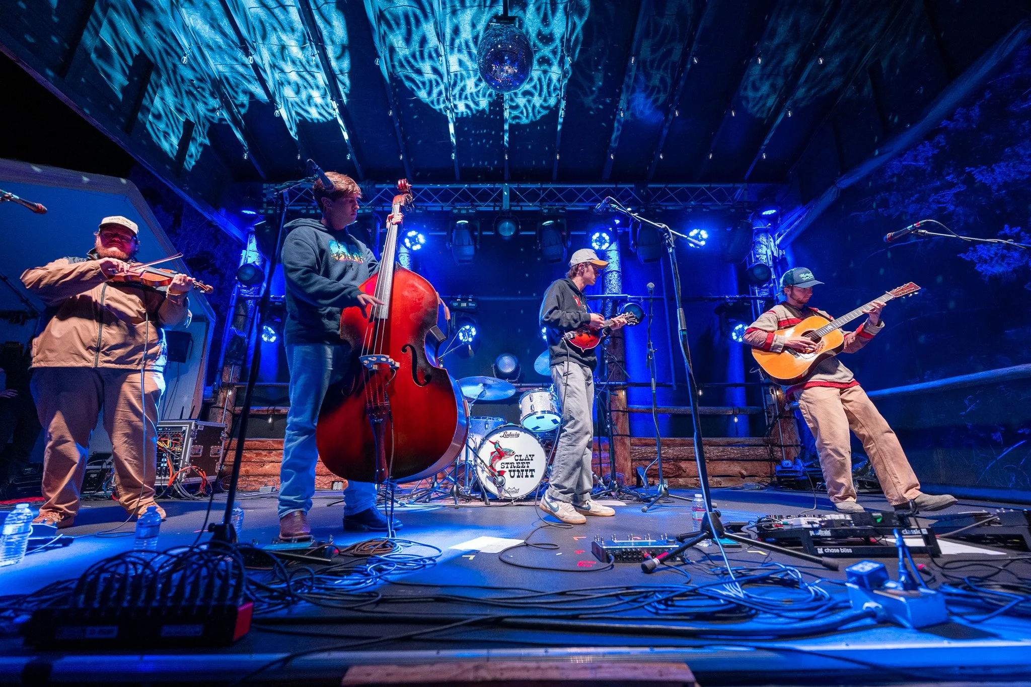 A band of five musicians performing on a stage with blue lighting and a disco ball overhead. The band includes a violinist, a double bassist, a guitarist, and a drummer, with musical equipment and microphones set up around them.