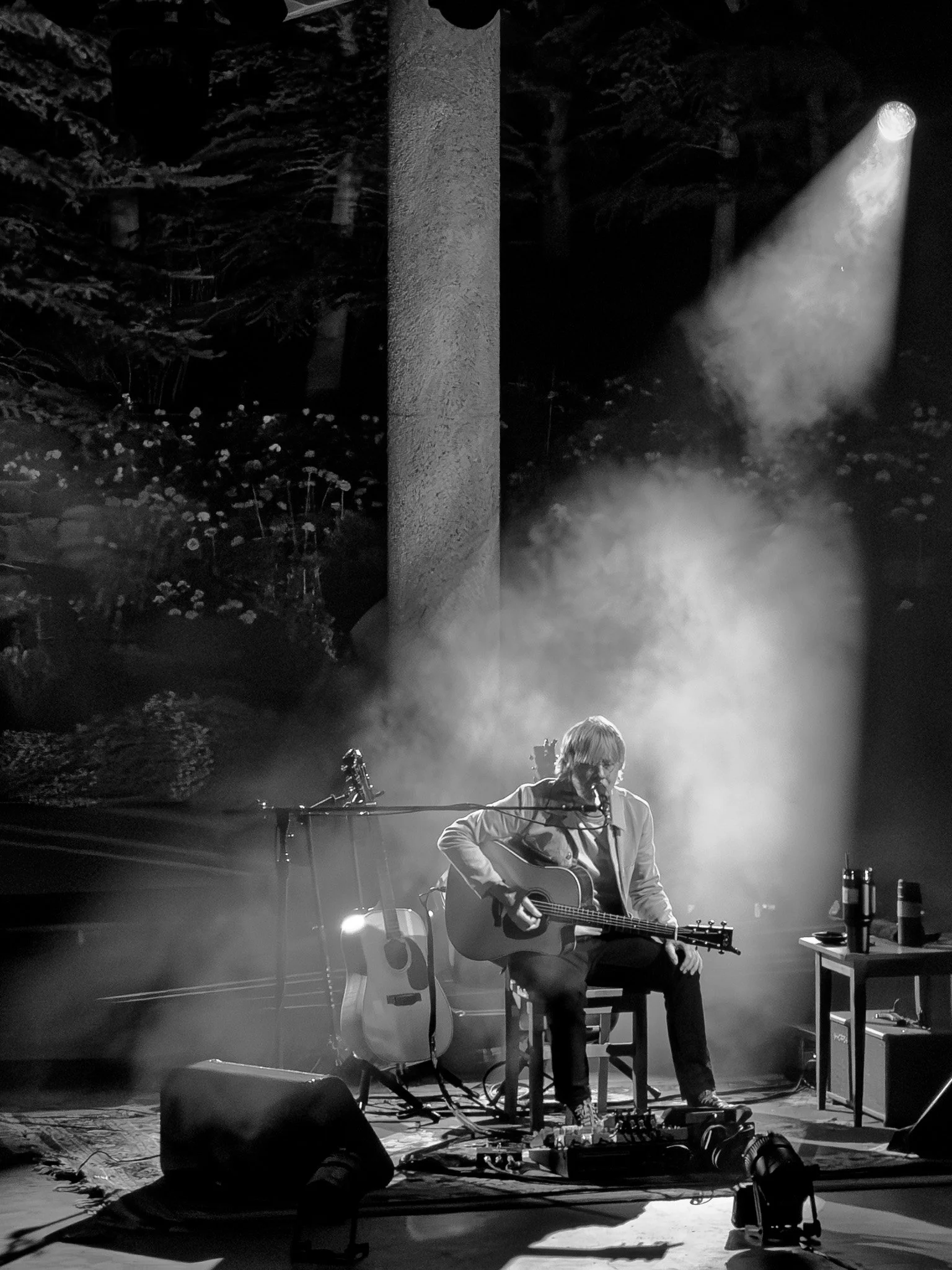 A man performing a solo guitar and vocals at a live music event, sitting on a chair with a microphone, in a dimly lit setting with stage lights and floral decorations in the background.