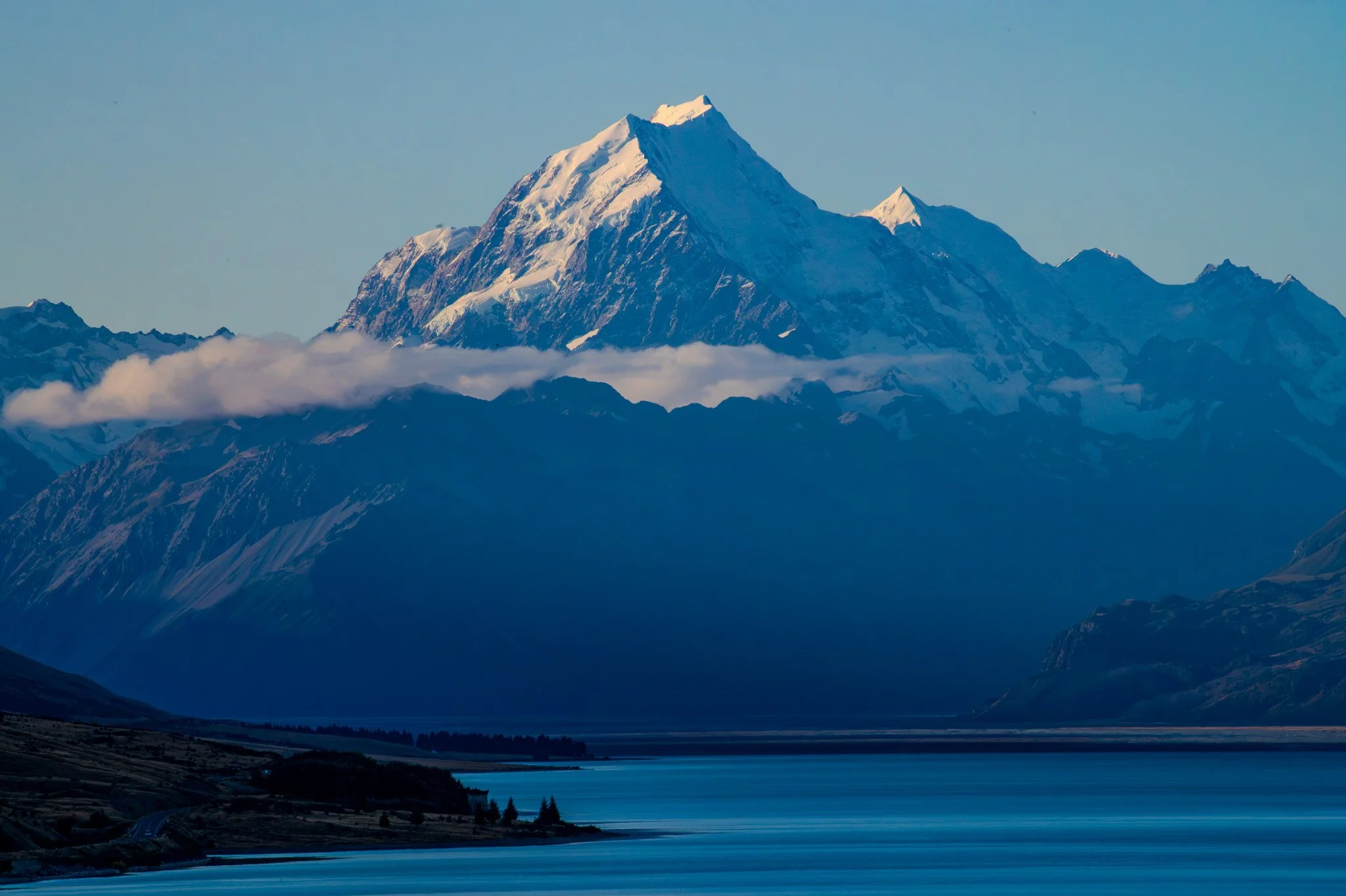 Snow-capped mountain range with a prominent peak, partially covered by clouds, near a lake with a forested shoreline in the foreground.