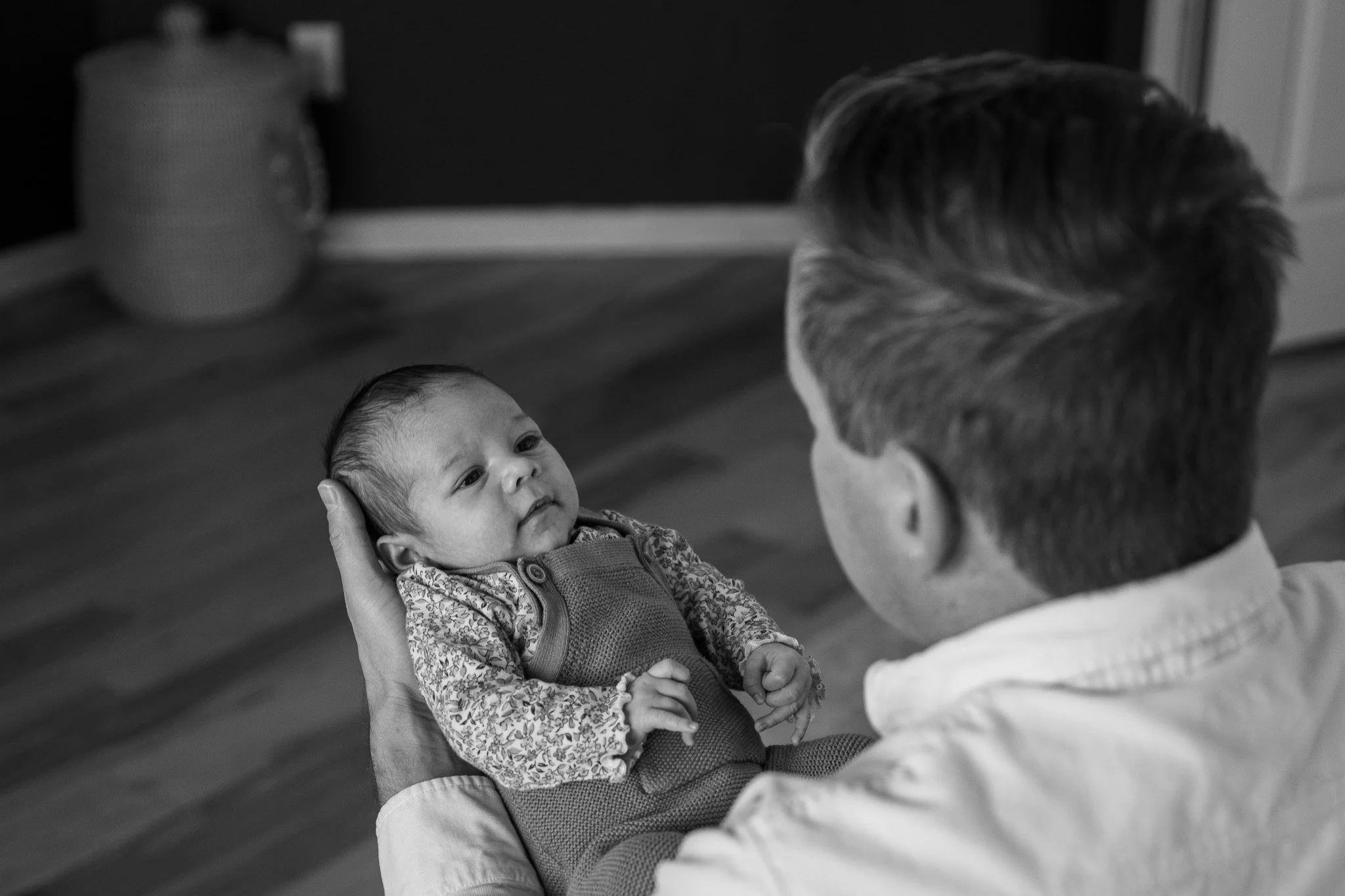 A person holding a baby in their hands, with the baby looking up at the person, who is facing them. The background includes a blurred dresser with a lamp.