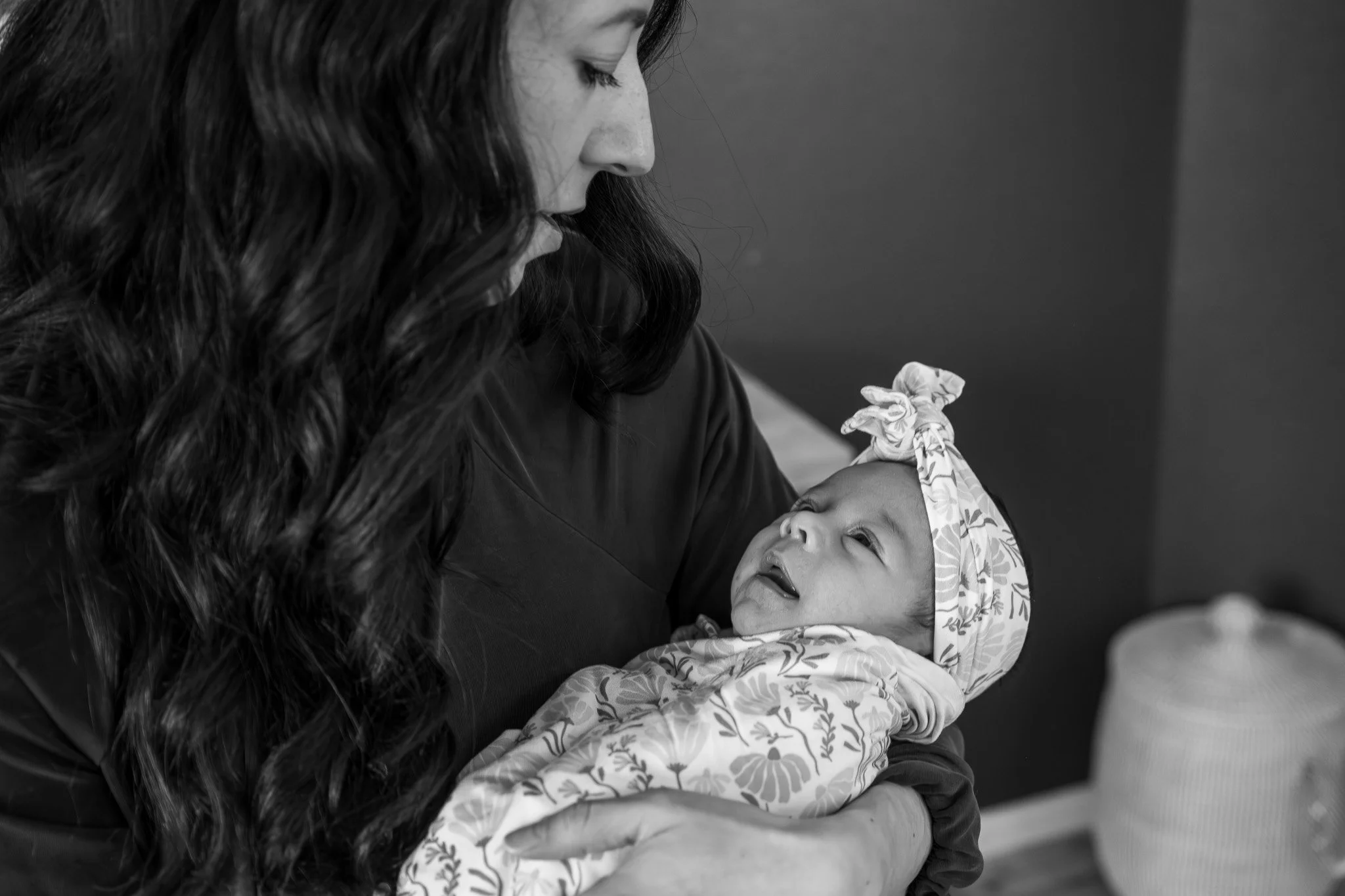 A woman with long dark hair holds a baby with a floral headband close to her face, both looking at each other affectionately in black and white.