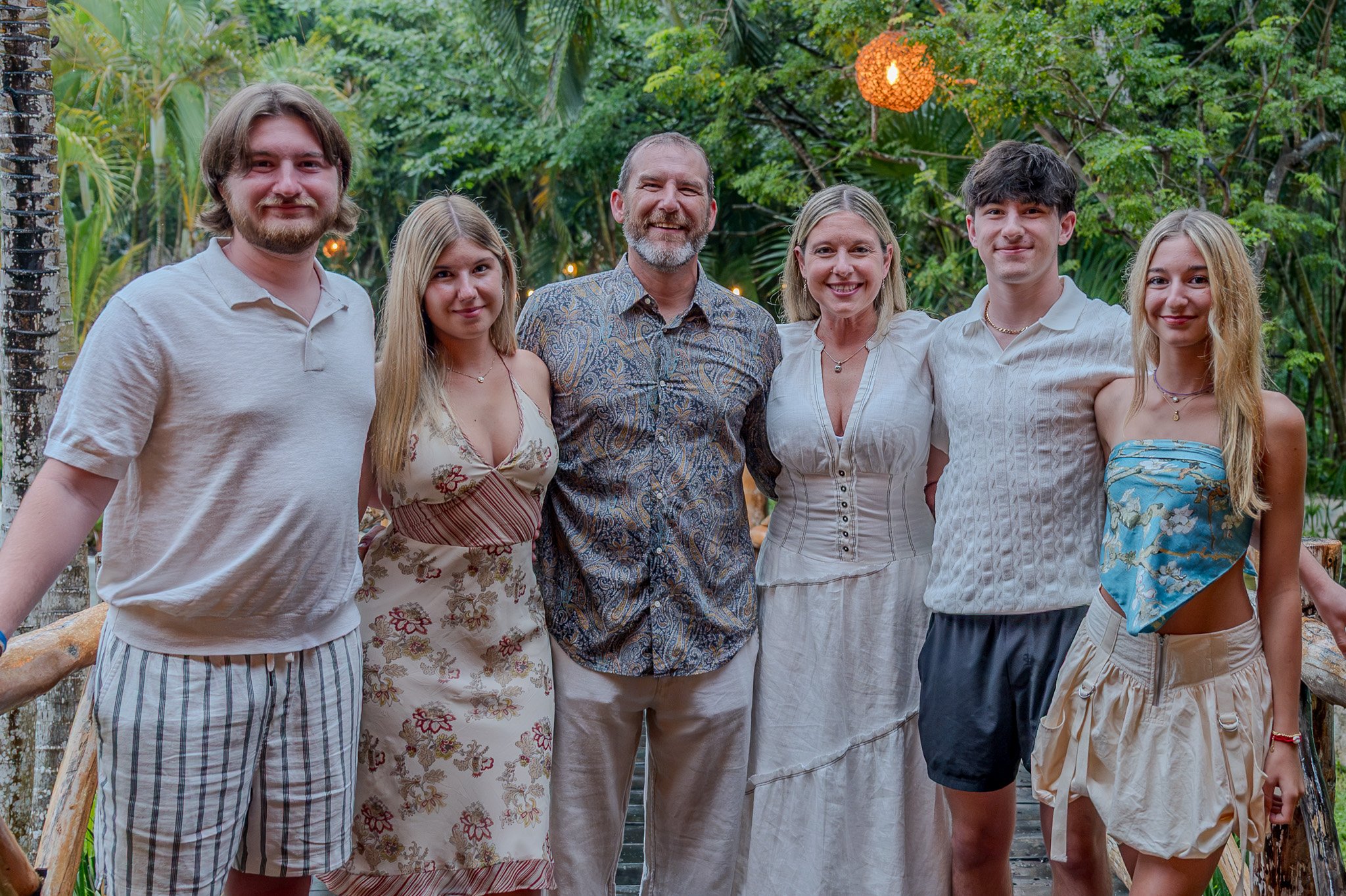 Group of seven people standing outdoors in a lush, green tropical setting with string lights and a hanging orange lantern.