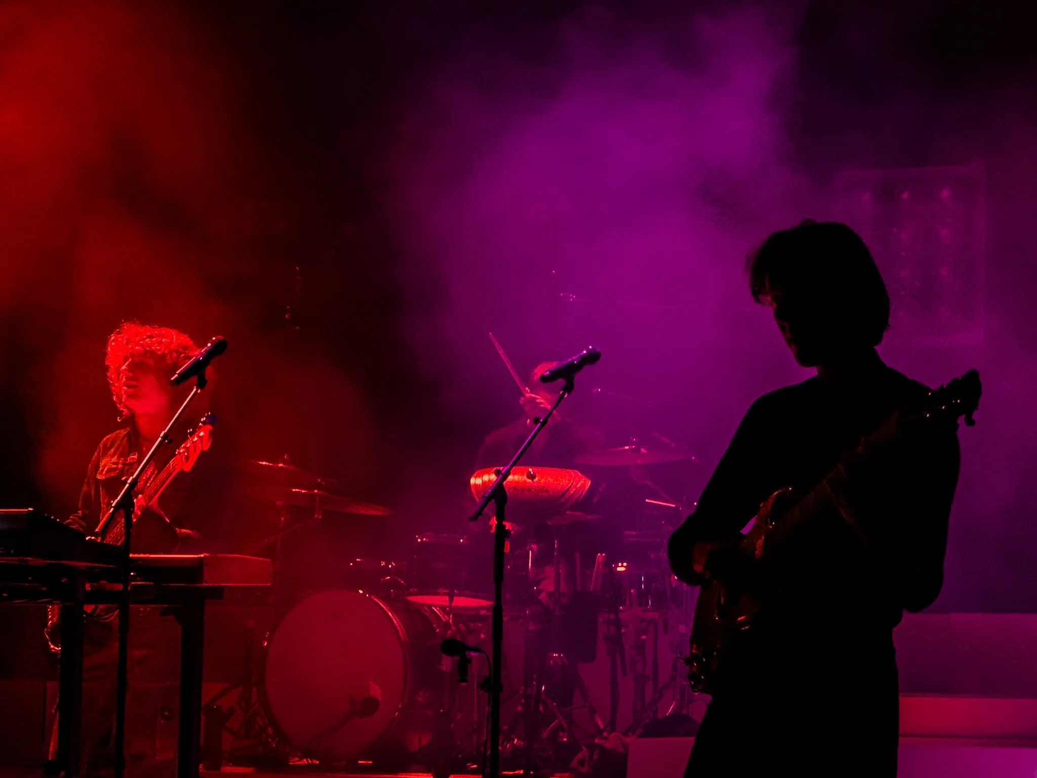 Silhouettes of musicians performing on stage with colorful red and purple stage lighting, musical instruments including a guitar, keyboard, drums, and microphones.