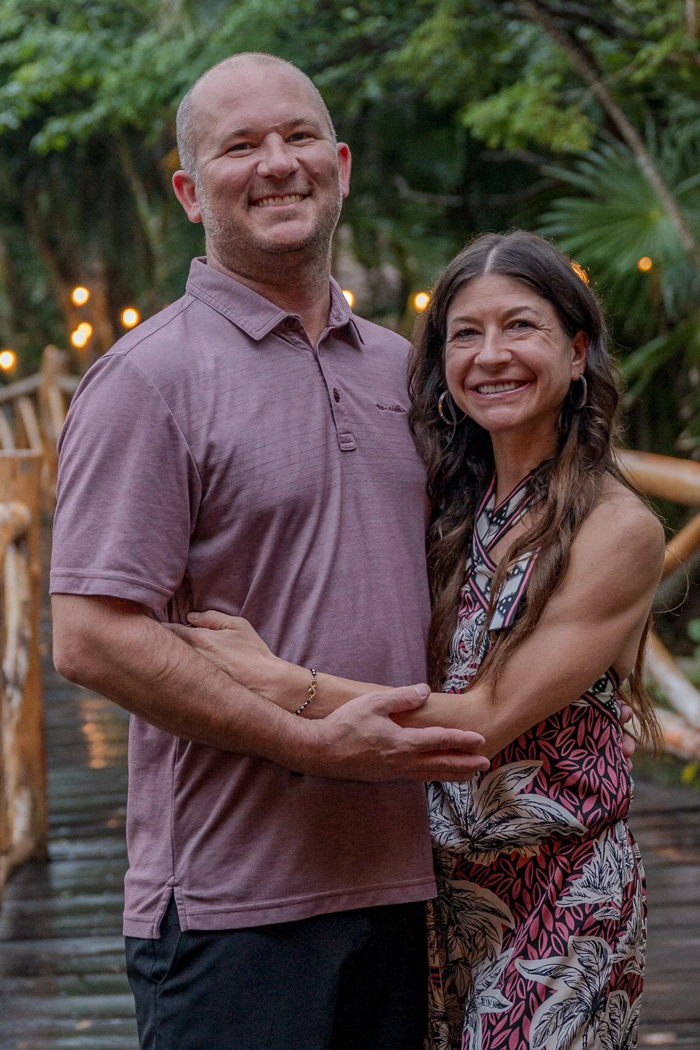 A smiling man and woman are embracing outdoors during evening, with trees and warm string lights in the background.