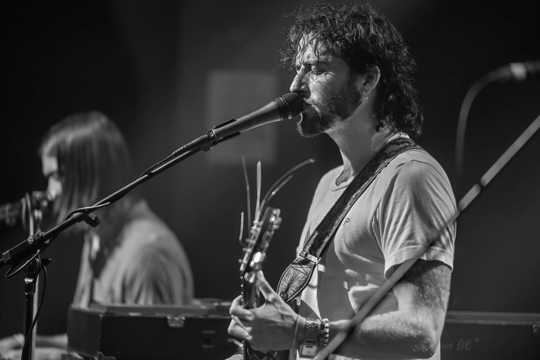 A black and white photo of a young man with curly hair and a beard singing into a microphone while playing an electric guitar, with another musician partially visible in the background.