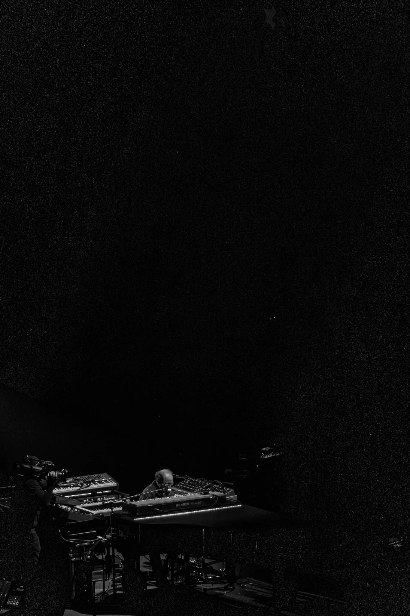 A black and white photograph of a music performance stage showing two musicians playing keyboard instruments, with one standing and the other seated, against a dark background.