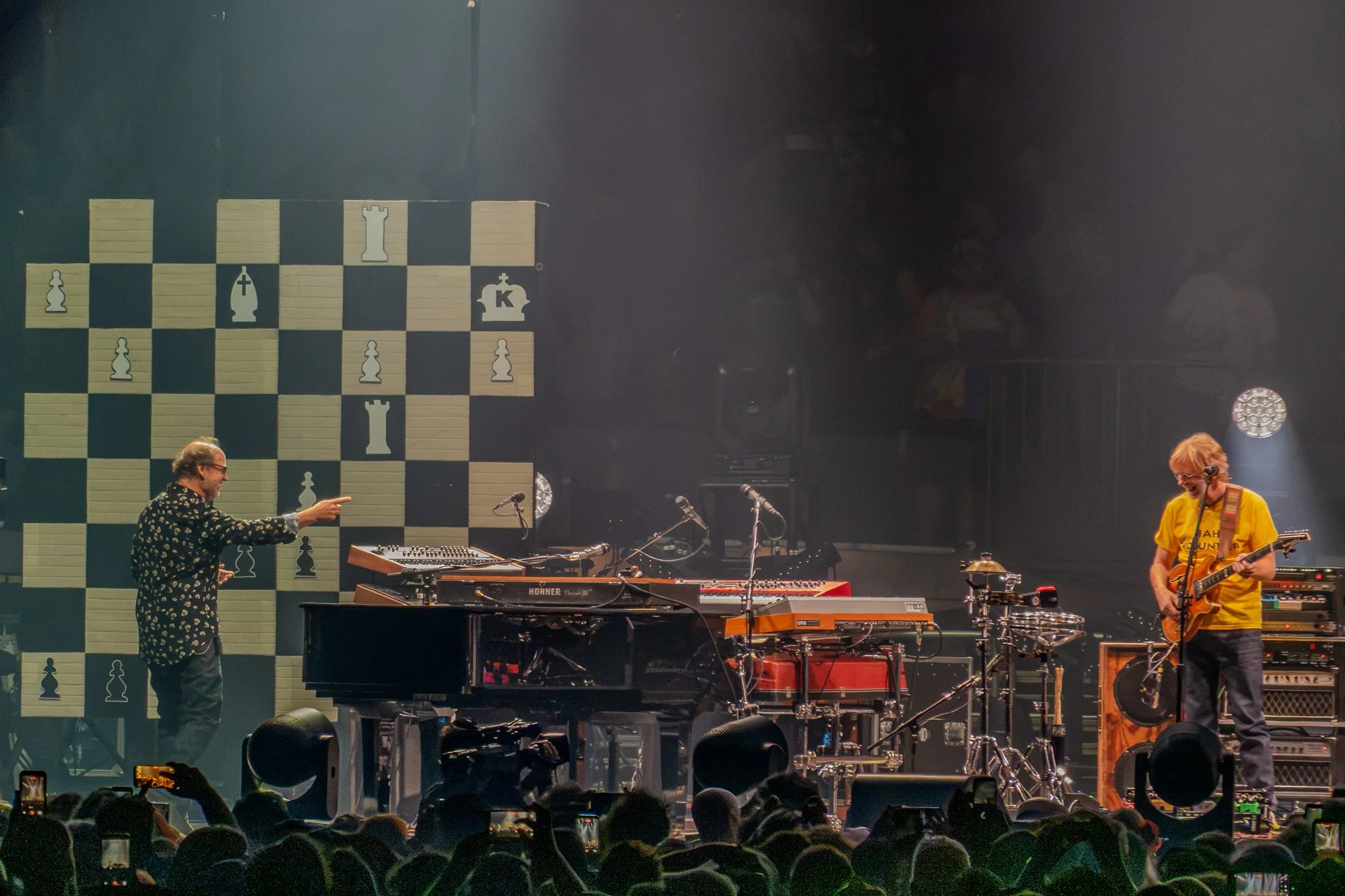 A live music performance on stage with two musicians, one playing the keyboard and the other playing the guitar, in front of a large chess-themed backdrop. The stage is filled with music equipment and a crowd of audience members taking photos.