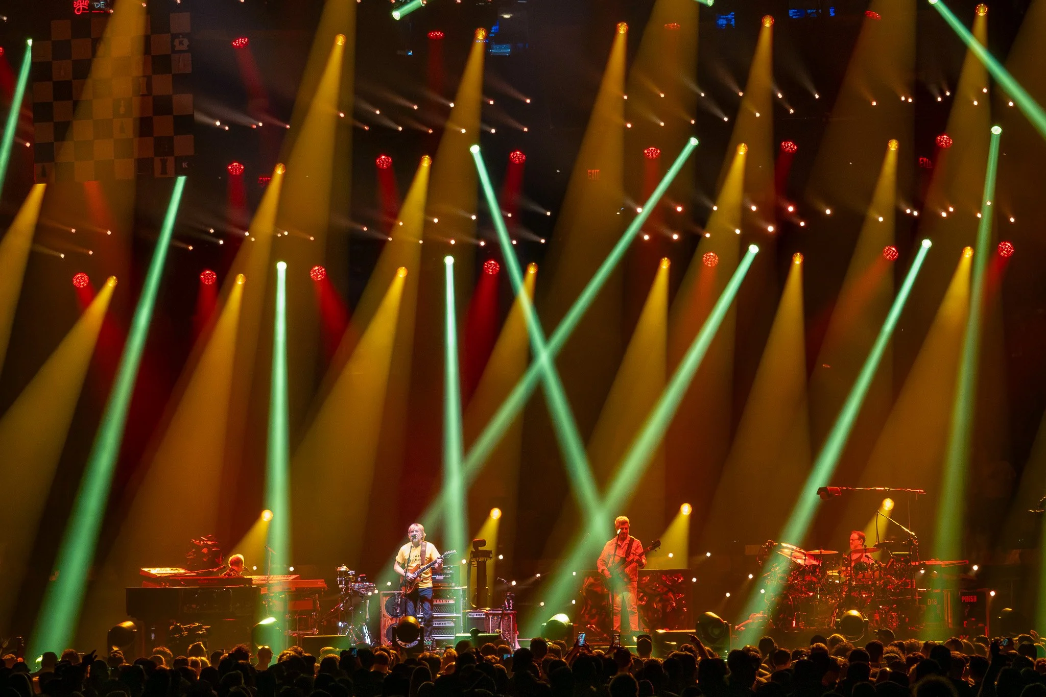 A live music concert with a band performing on stage. The stage is lit with colorful yellow, green, and red spotlights, and there is a large crowd in front. The band members include musicians playing guitars, drums, and keyboards. The background feat