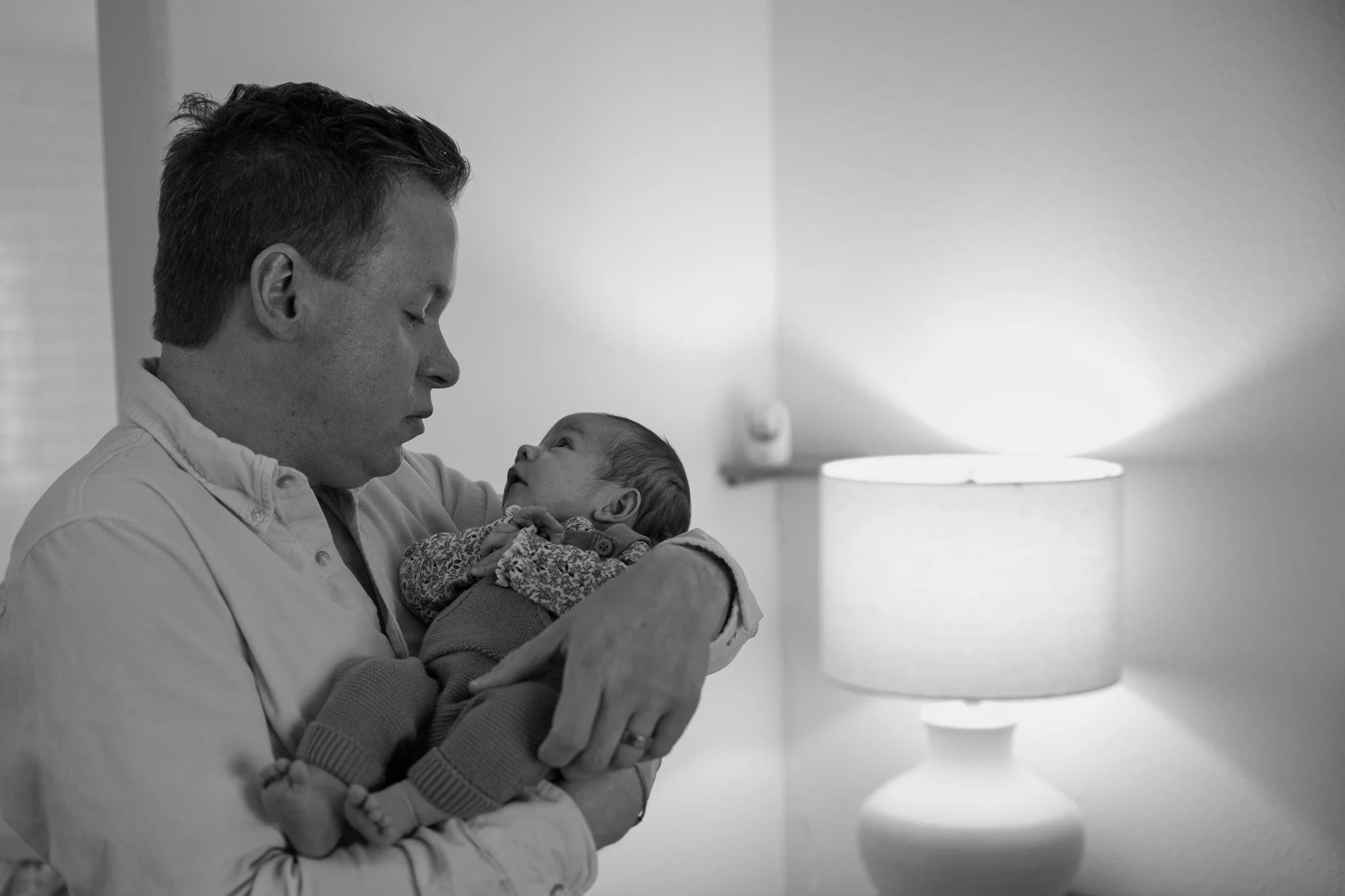 A man holding a baby close, looking at the baby with affection in a room with a modern table lamp in the background.