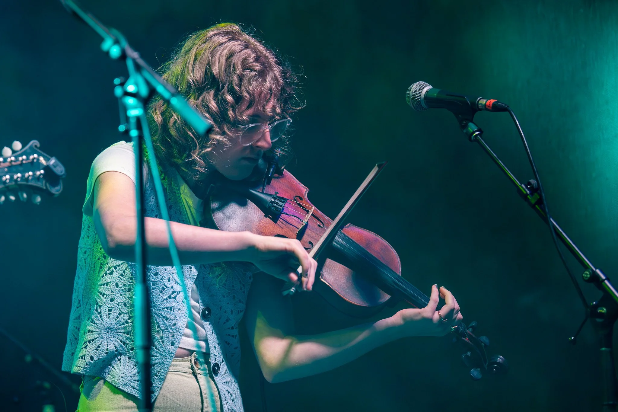 A woman with curly hair and glasses playing a violin on stage, with microphone and musical equipment around her, illuminated by stage lighting.