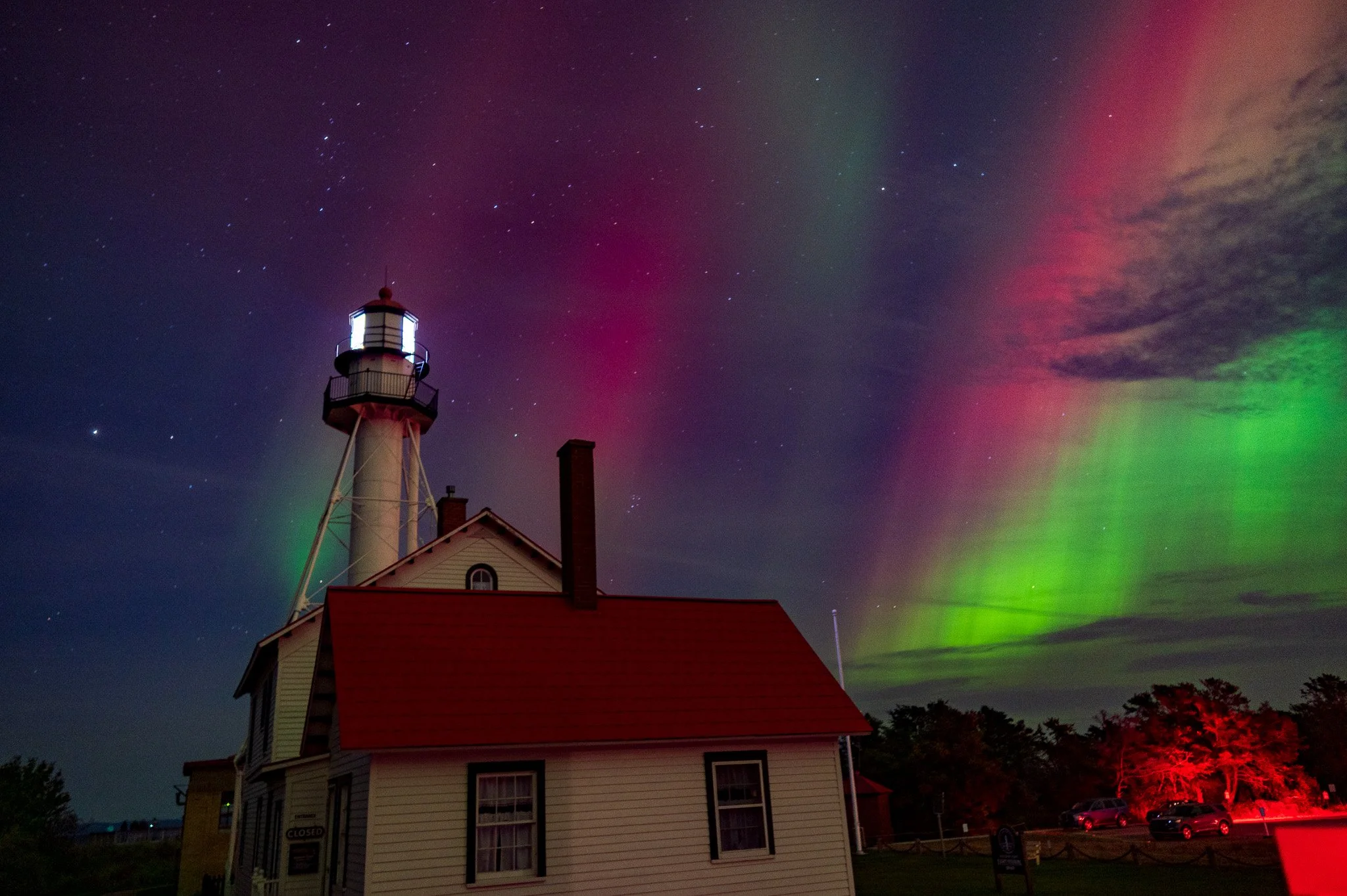 A lighthouse building with a red roof seen at night under a colorful northern lights sky with stars.