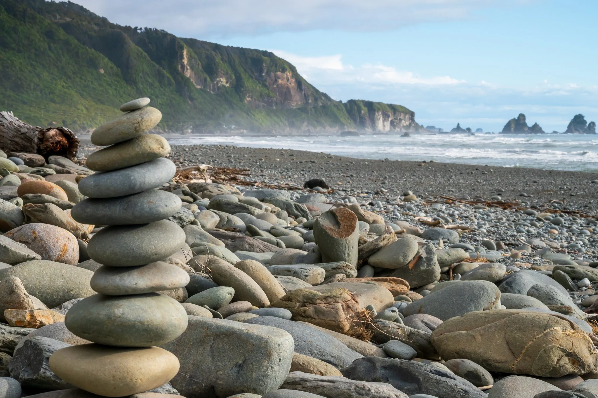 A stack of smooth, rounded rocks on a pebble-covered beach with green cliffs and ocean waves in the background.