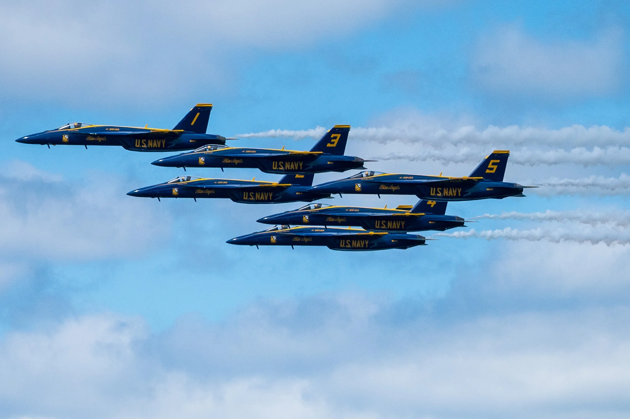 A formation of five U.S. Navy Blue Angels fighter jets flying in a diagonal line with blue sky and clouds in the background.