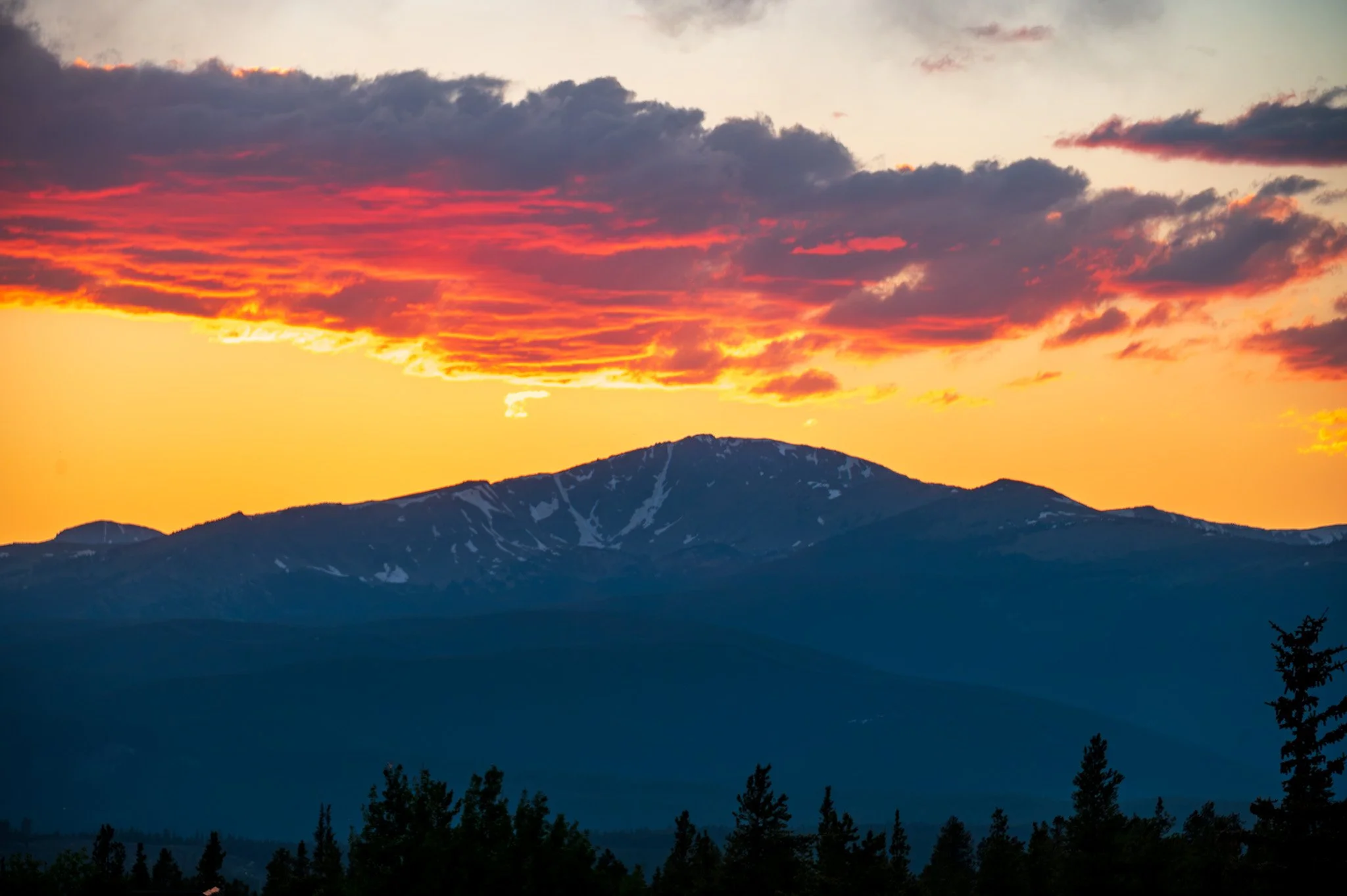 A mountain range at sunset with a colorful sky of orange, pink, and purple hues, and dark silhouetted trees in the foreground.