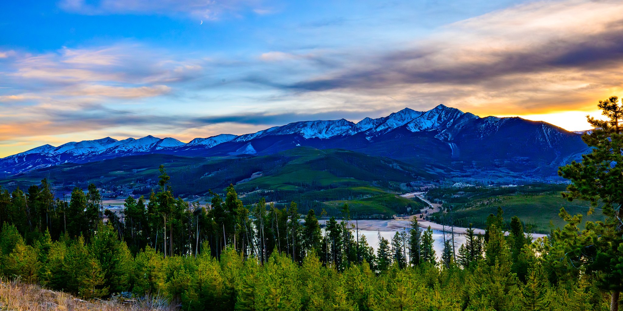 A scenic view of snow-capped mountains at sunset with a colorful sky, green forest, and a small river or lake in the foreground.