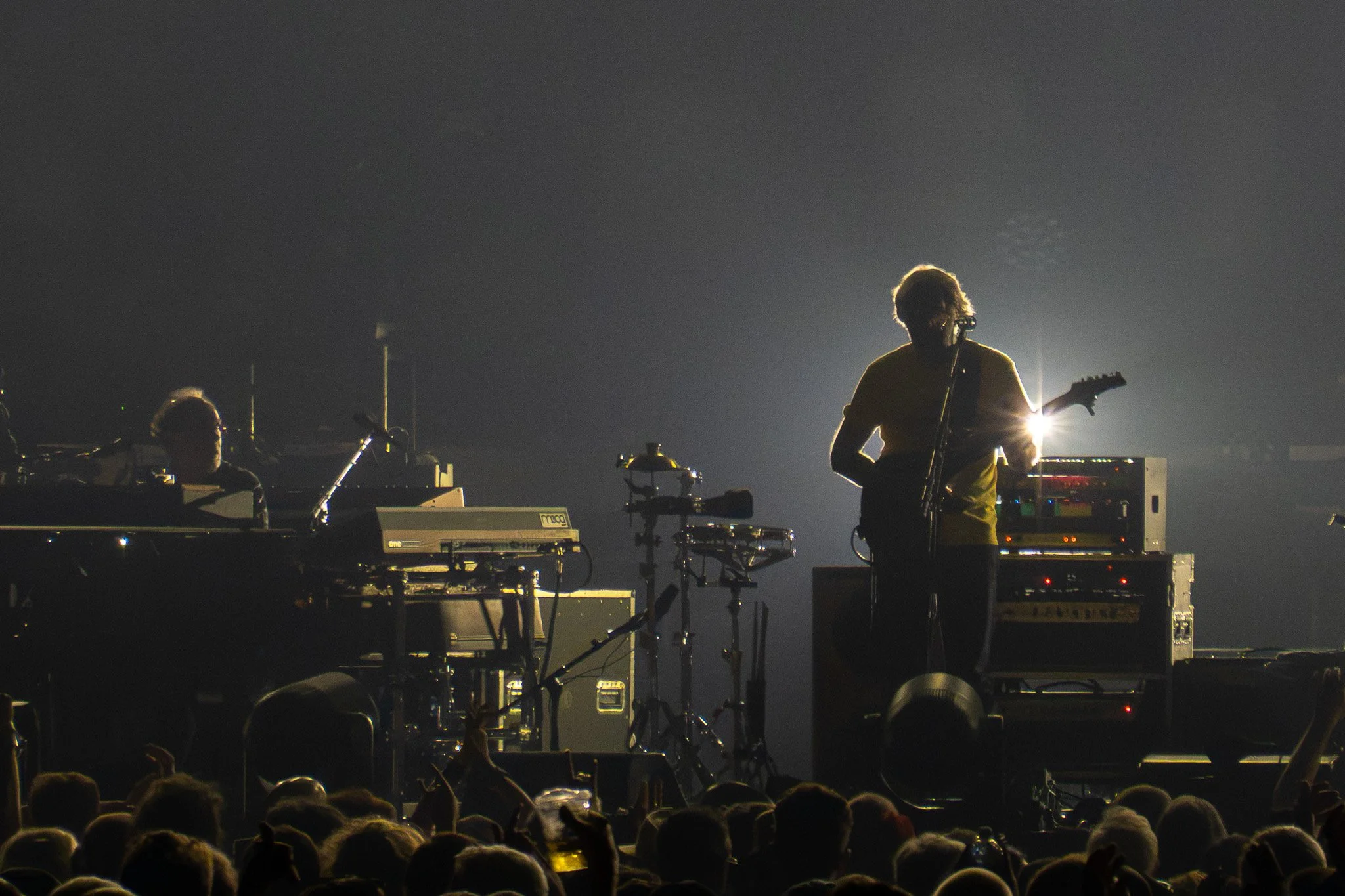 A silhouette of a musician playing guitar on stage with bright light behind, accompanied by a drummer, during a concert at night.