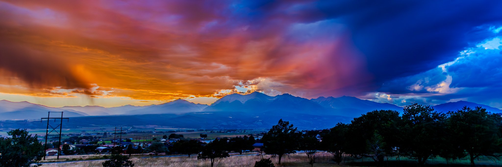 Mountains at sunset with colorful orange, purple, and blue sky, trees in the foreground, and power lines.