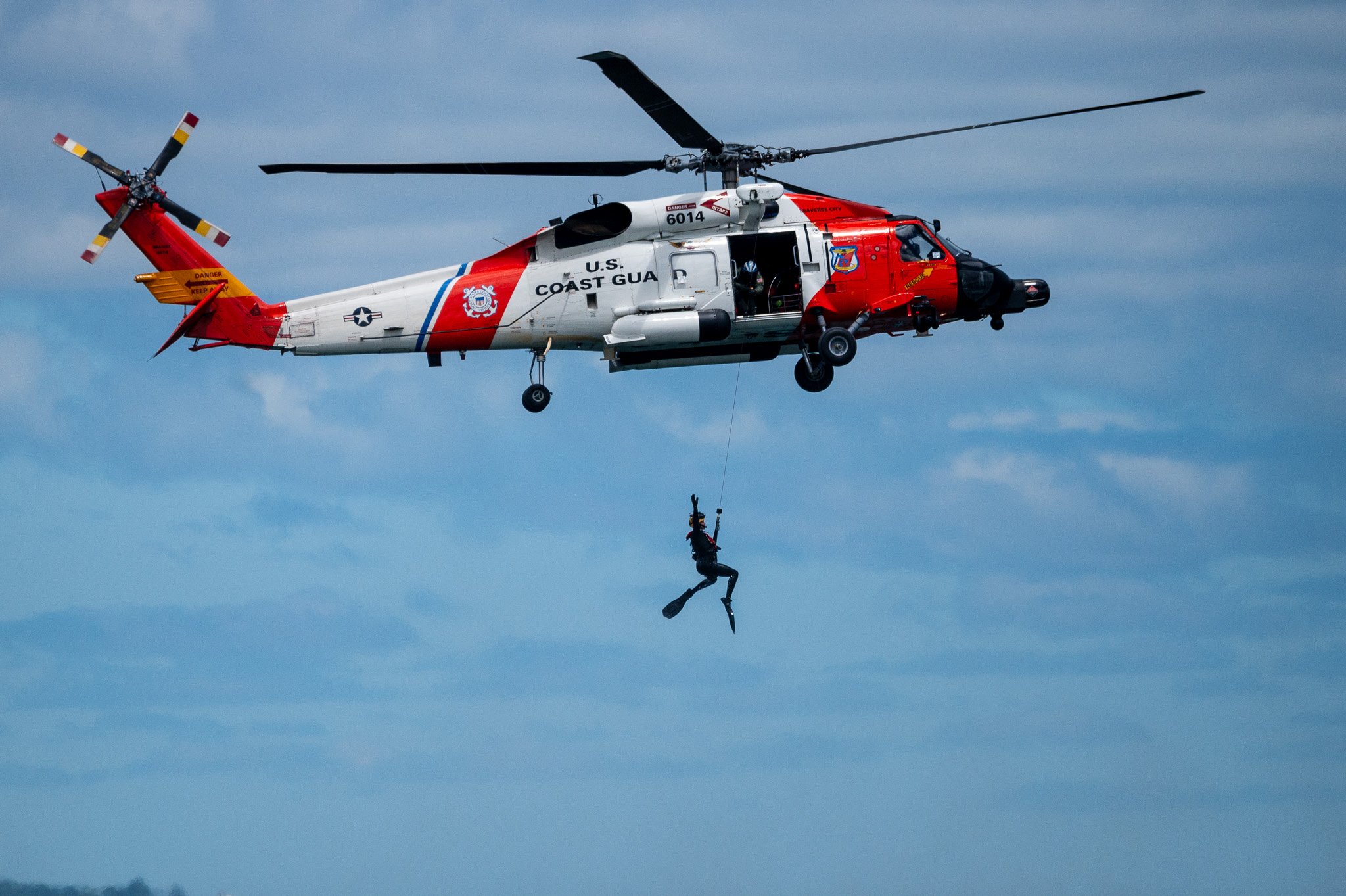U.S. Coast Guard helicopter in flight with a rescue diver hanging beneath it, against a backdrop of blue sky and clouds.
