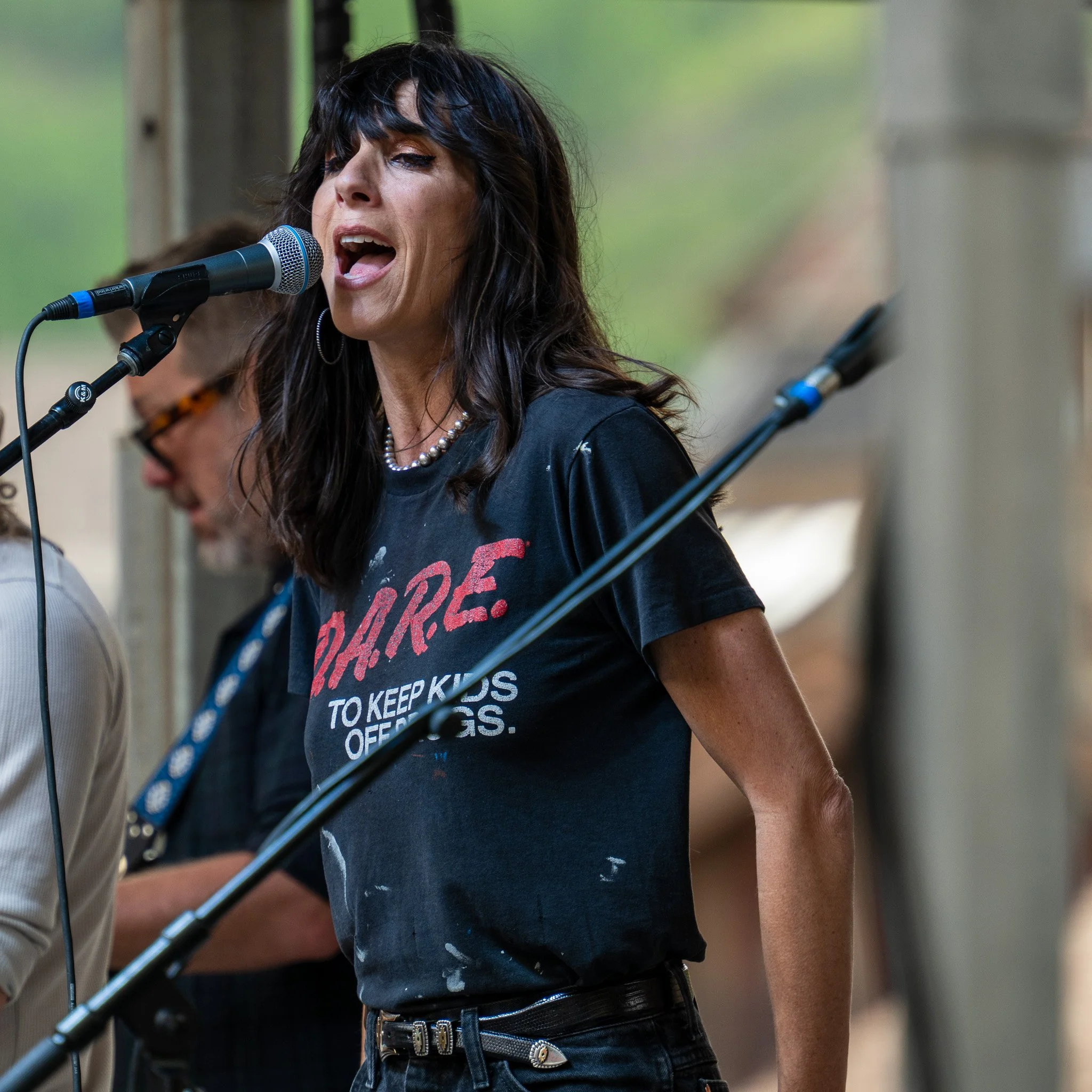 A woman with black hair and bangs singing into a microphone at an outdoor event.