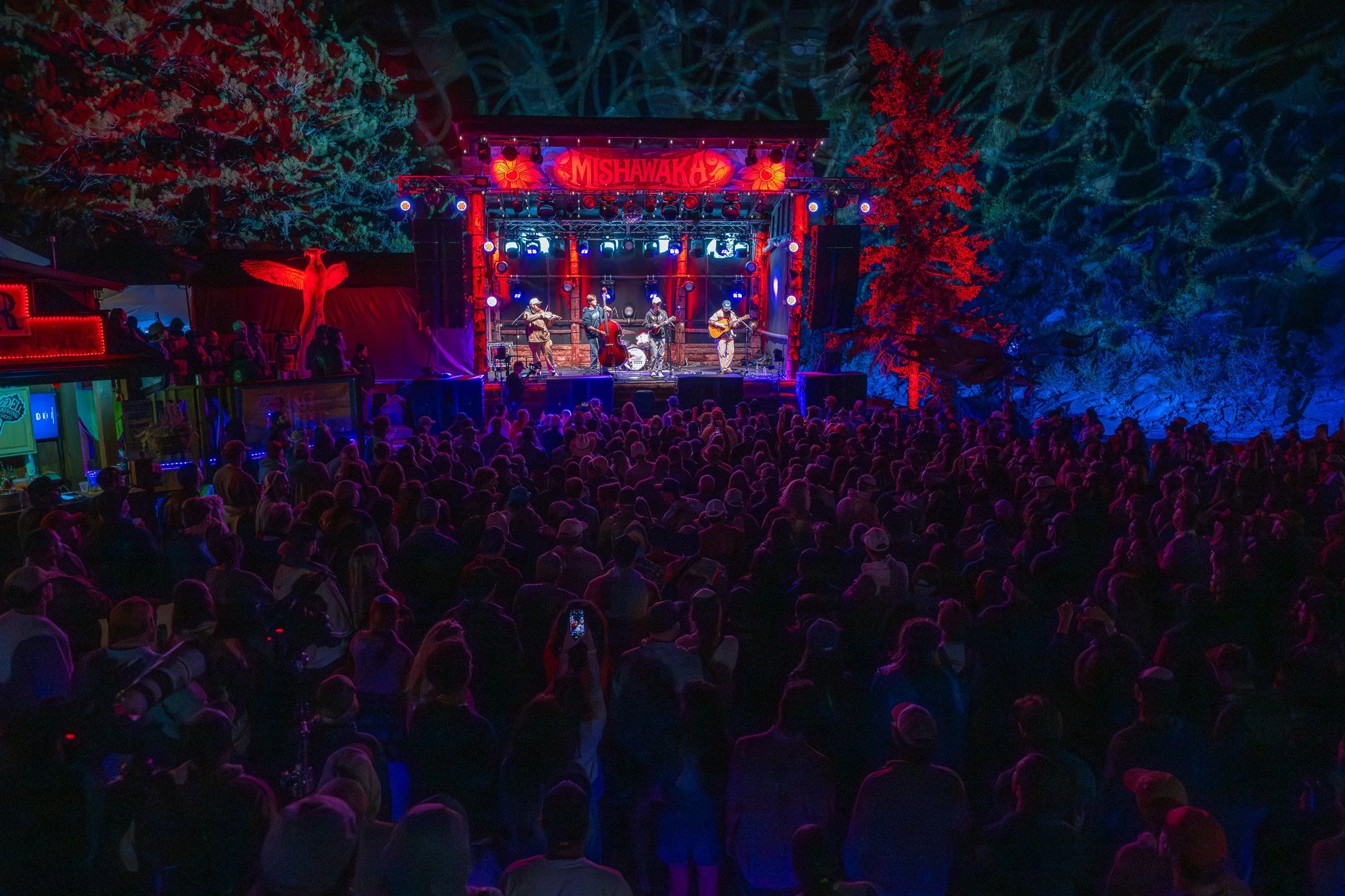 An outdoor stage with a band performing at night, decorated with colorful lights and signage reading 'Mishawaka,' surrounded by a large crowd of people and tall trees.