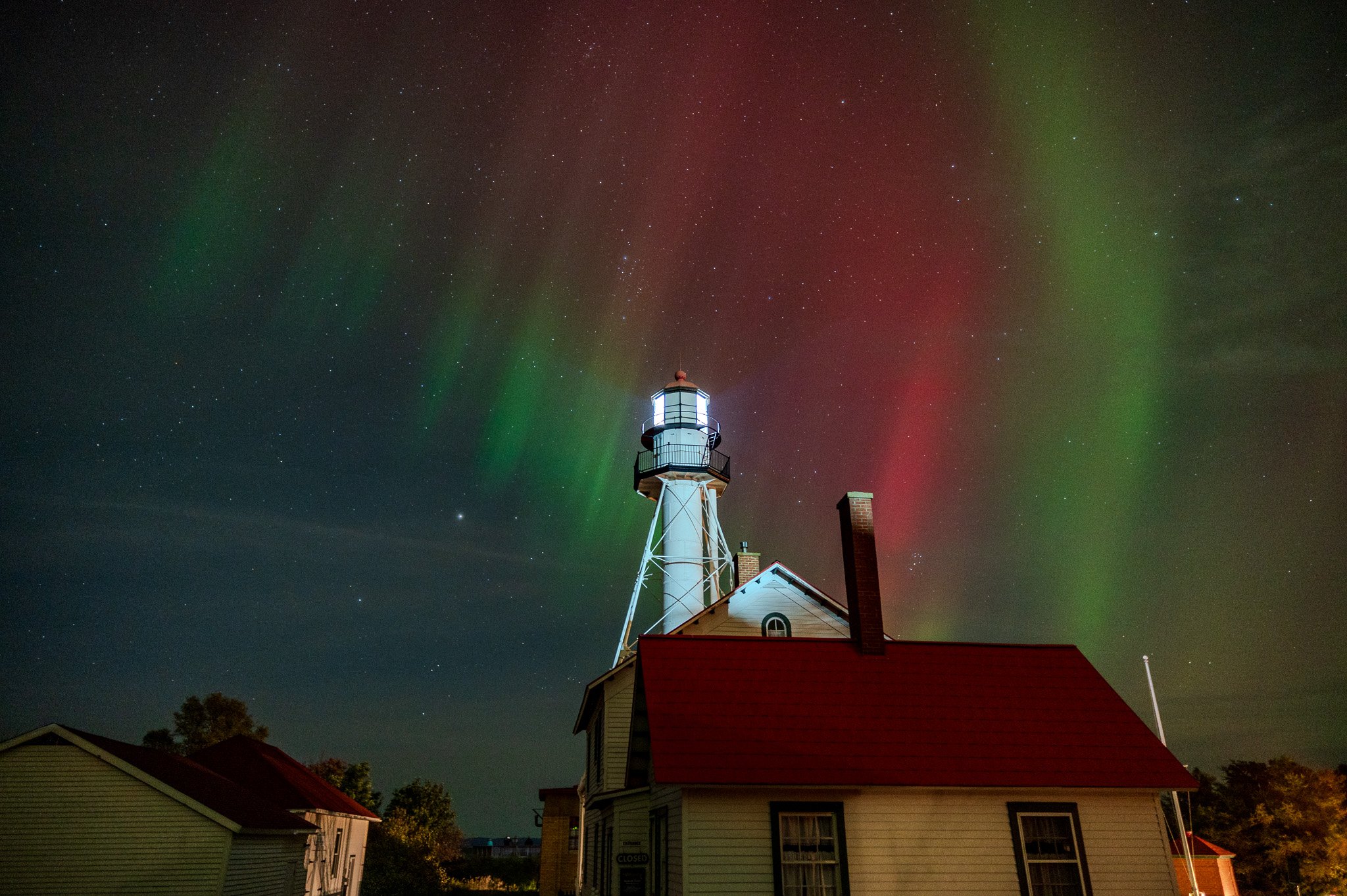 Night sky with stars and colorful aurora borealis over a lighthouse and houses.