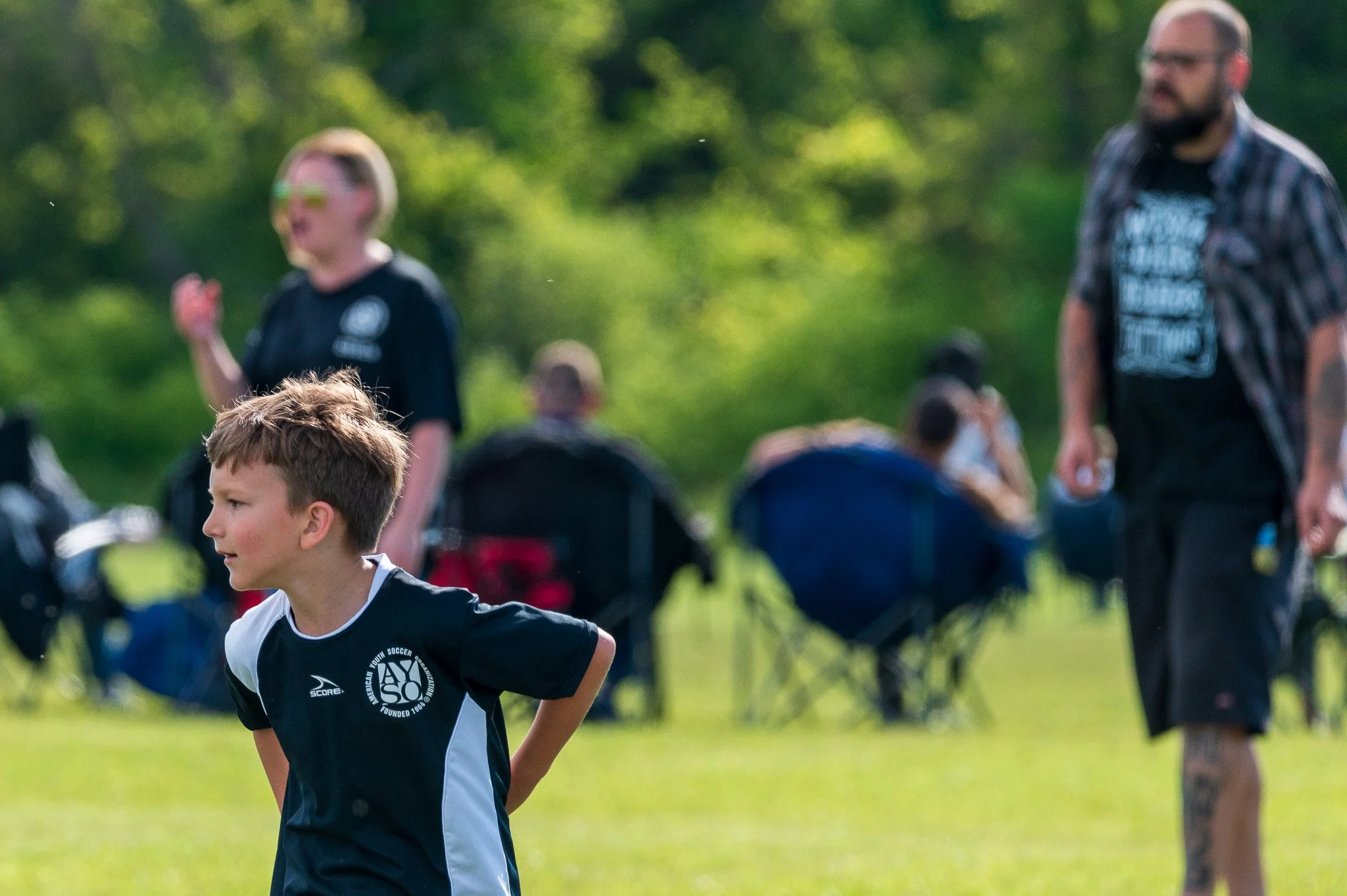 A young boy in a black and white soccer jersey outdoors during daylight, with a group of people, mostly children, in the background. The setting appears to be a park or a field with green trees.