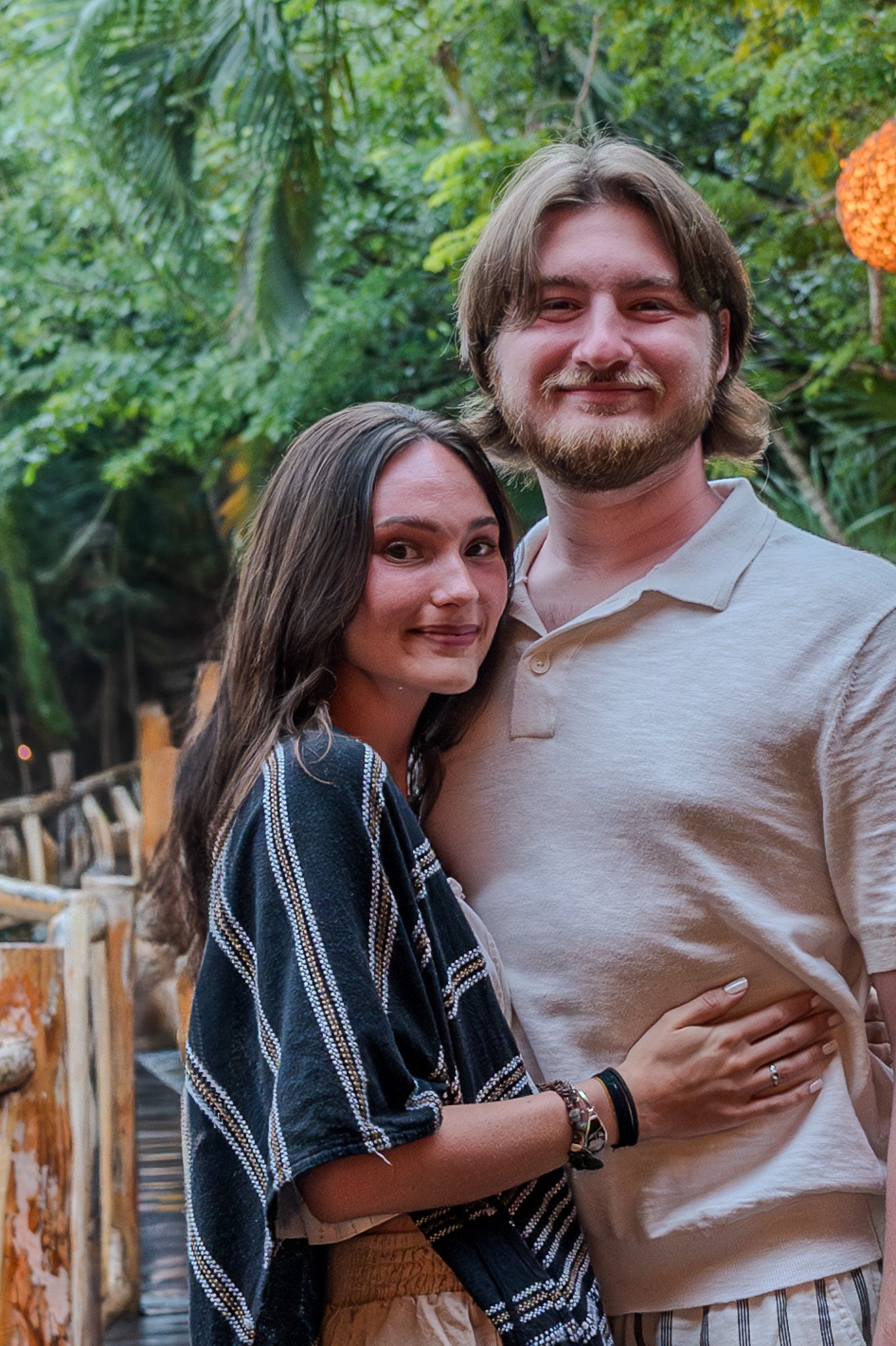 A young woman and man standing close together outdoors in a lush, green environment.