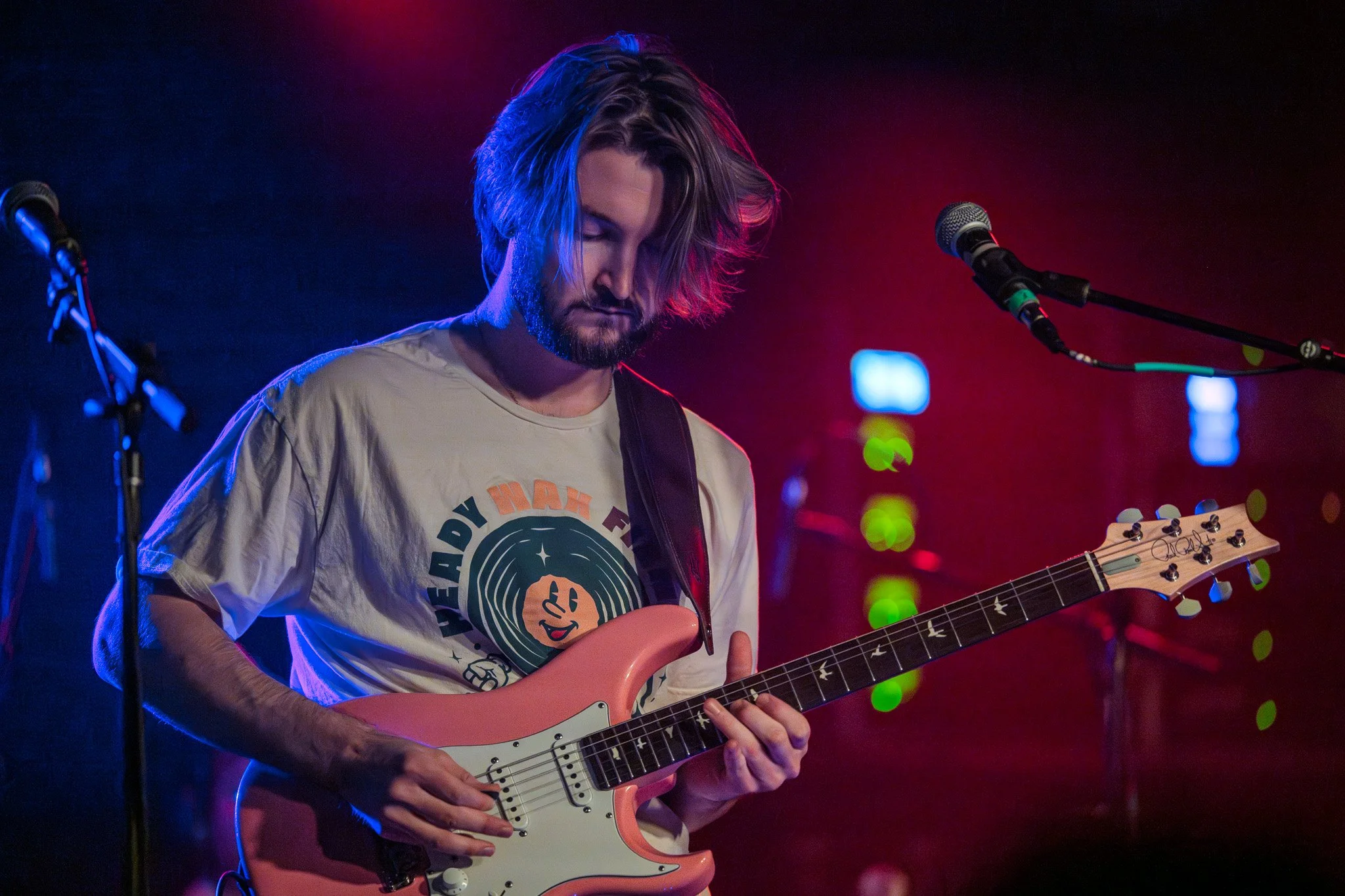 A male musician with shoulder-length hair and a beard playing a pink electric guitar on stage, with colorful stage lighting.
