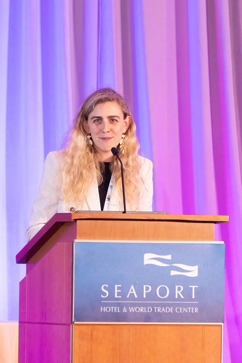A woman with long blonde hair wearing earrings and a cream-colored blazer stands at a podium with a microphone, speaking at a conference. The podium has a sign that says "Seaport Hotel & World Trade Center." The background features purple and pink cu