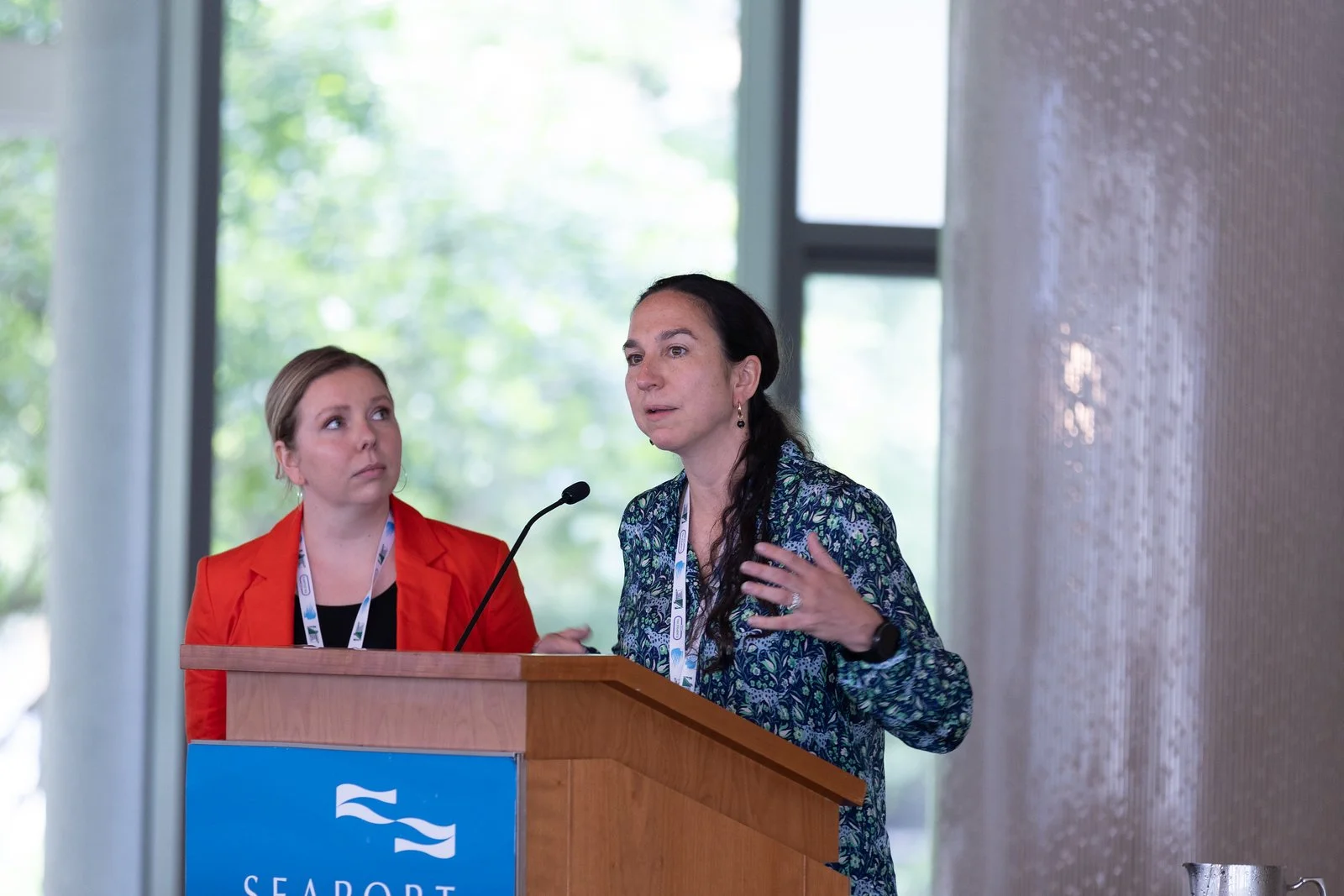 Two women at a conference, one speaking at a podium and the other listening, with a background of large windows and natural light.