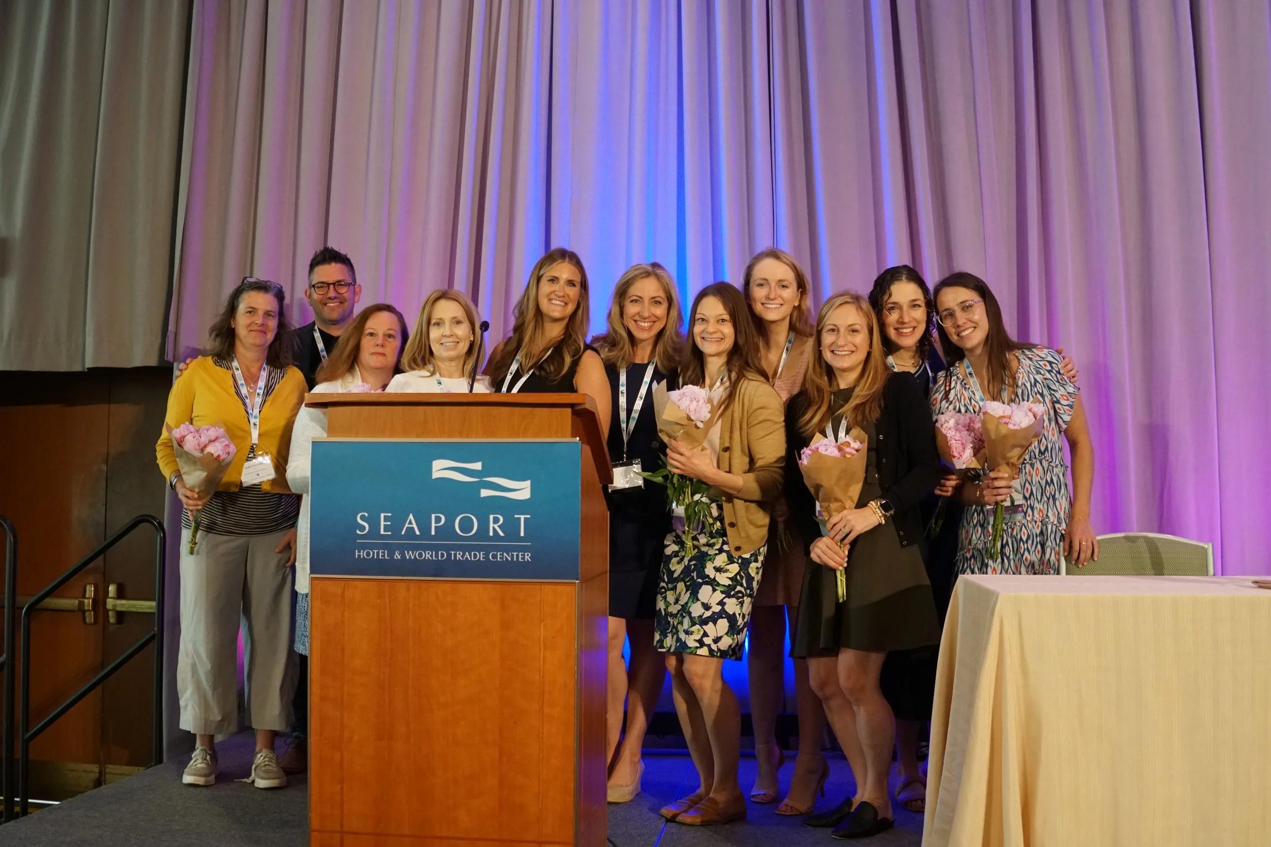 Group of women and one man standing together on stage at a conference, smiling, with some holding bouquets, behind a podium with a blue sign for Seaport Hotel & World Trade Center, in front of purple curtains.