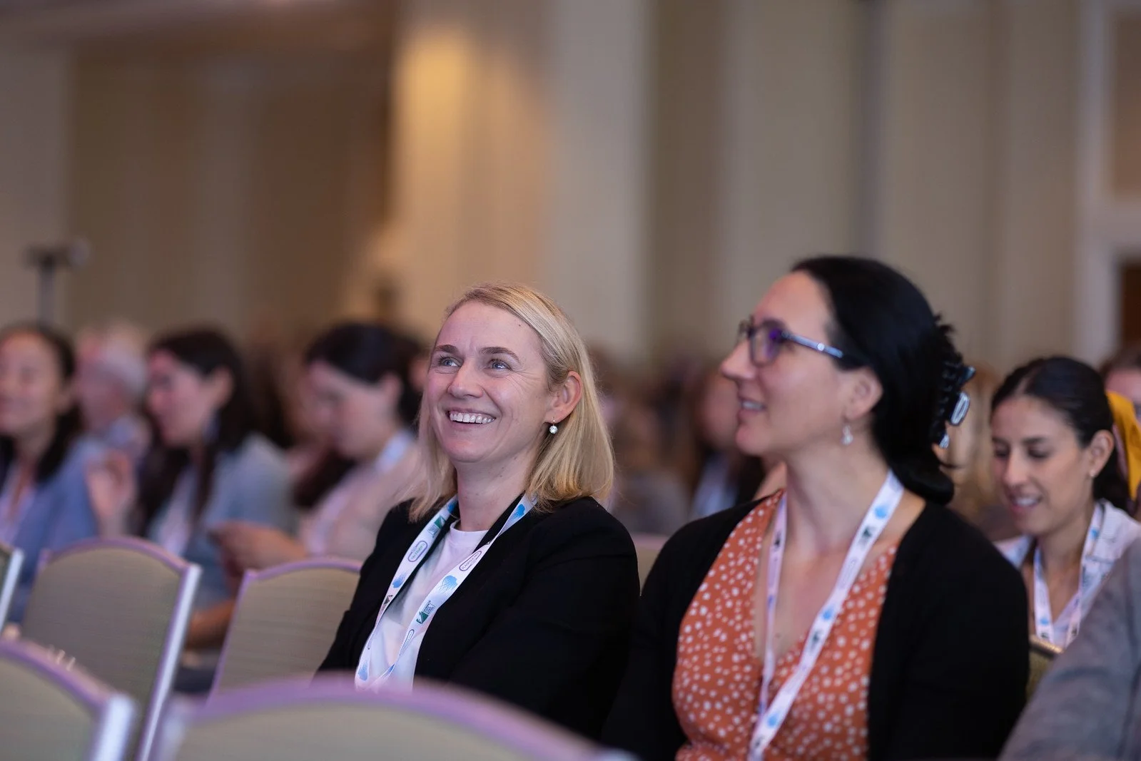 Multiple women attending a conference, sitting in chairs, wearing conference badges and smiling, with a blurred background of other attendees.